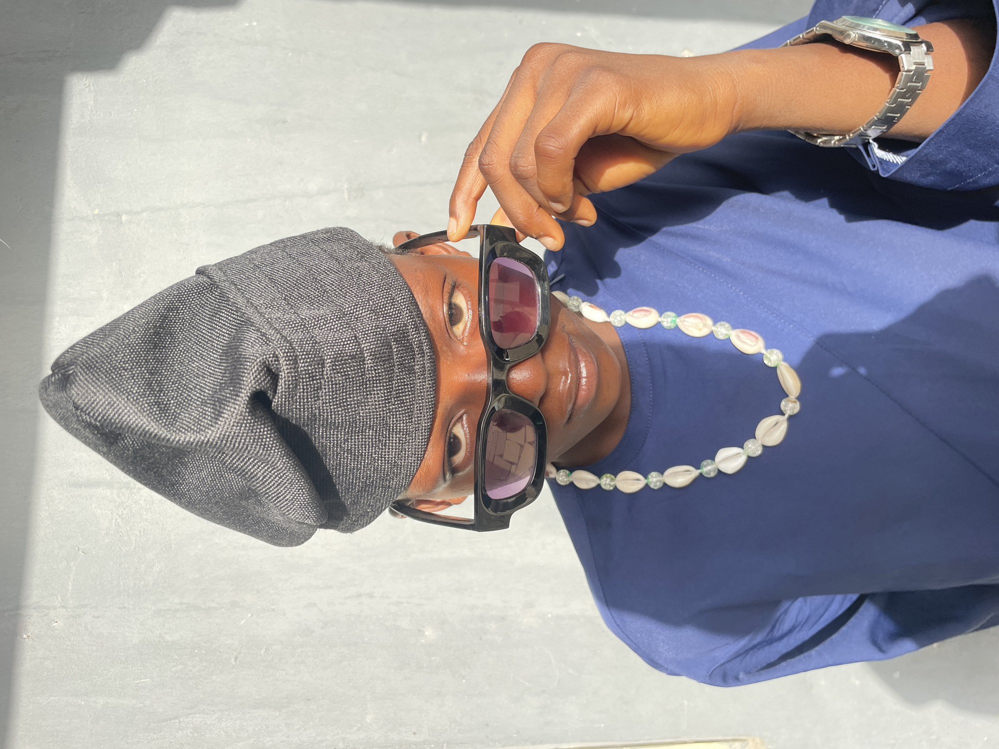 An eye-level portrait shot captures a person, likely male, against a bright, light-colored concrete or paved surface, with the image rotated 90 degrees counter-clockwise. The individual has dark skin and is looking upwards relative to the image frame, with a neutral to slightly serious expression.
They are wearing a dark grey, textured cap or beanie that covers their head. A pair of stylish, black-framed rectangular sunglasses with dark, possibly purple-tinted lenses are perched on their face. Their right hand is visible, with dark skin, gently holding the frame of the sunglasses, pulling them slightly down, revealing their right eye. The person is adorned with a distinctive necklace made of light-colored cowrie shells interspersed with translucent, light green beads.
They are dressed in a plain blue collared shirt or tunic. On their left wrist, a silver-toned metal watch with a link bracelet is visible. Strong, defined shadows are cast on the blue shirt and the background surface, indicating bright, direct sunlight. The background is simple and uncluttered, suggesting an outdoor setting under a clear sky.
No text is visible in the image. The setting is consistent with warm, sunny weather in Badagry, Nigeria.