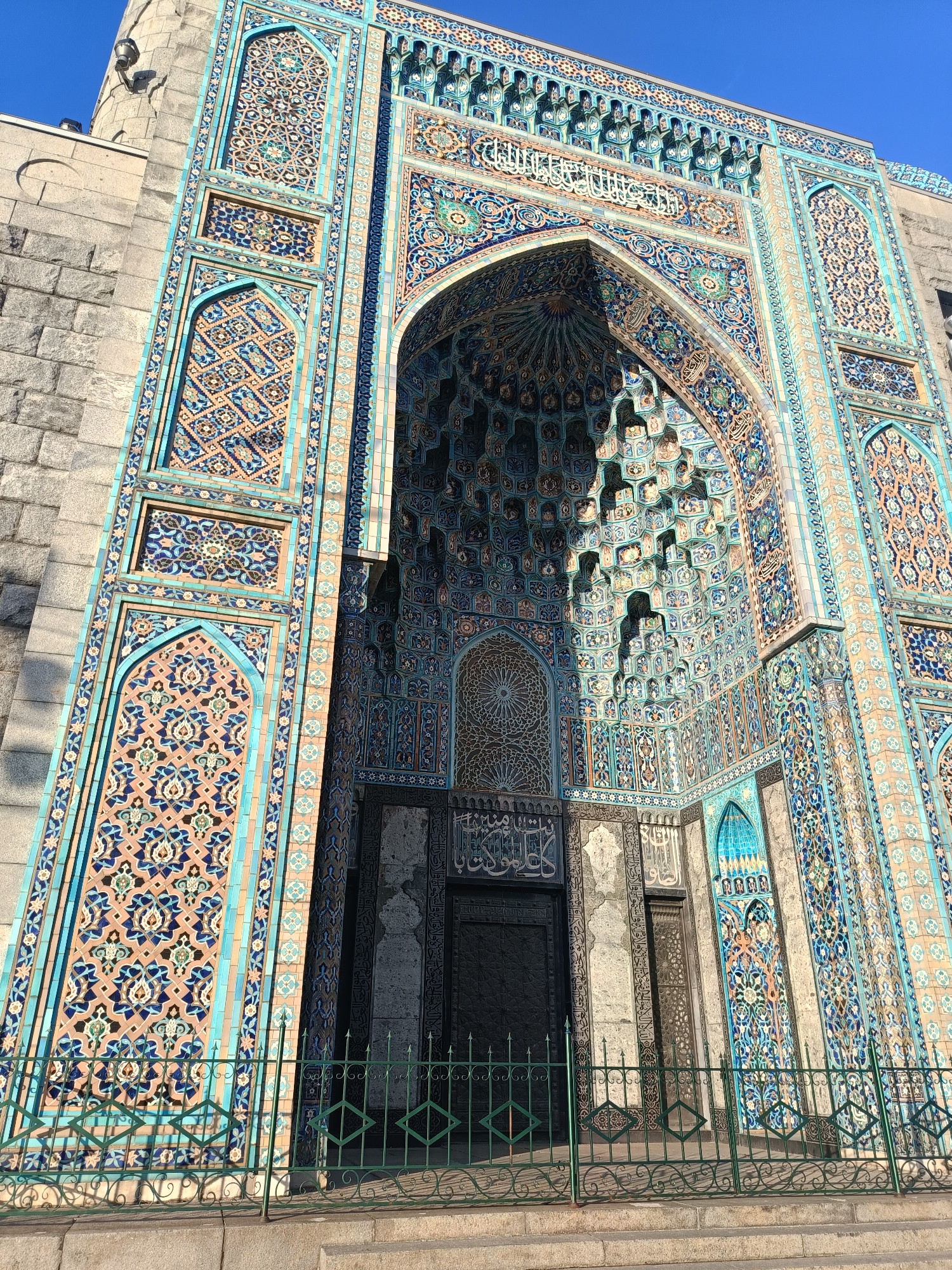 The image shows the exterior of the Saint Petersburg Mosque, located in Saint Petersburg, Russia. The mosque features a prominent, ornately decorated entrance with intricate mosaic tilework in shades of blue, turquoise, and peach. The tiles form geometric patterns and floral motifs. Above the entrance, Arabic calligraphy is visible. The main structure of the mosque is made of large, gray stone blocks, contrasting with the colorful tilework. A green metal fence with decorative scrollwork and diamond shapes stands in front of the mosque, separating it from a stone walkway and stairs. The sky above is a clear, bright blue. There are no people visible in the image. The perspective is from a low angle, looking upwards at the mosque's facade.