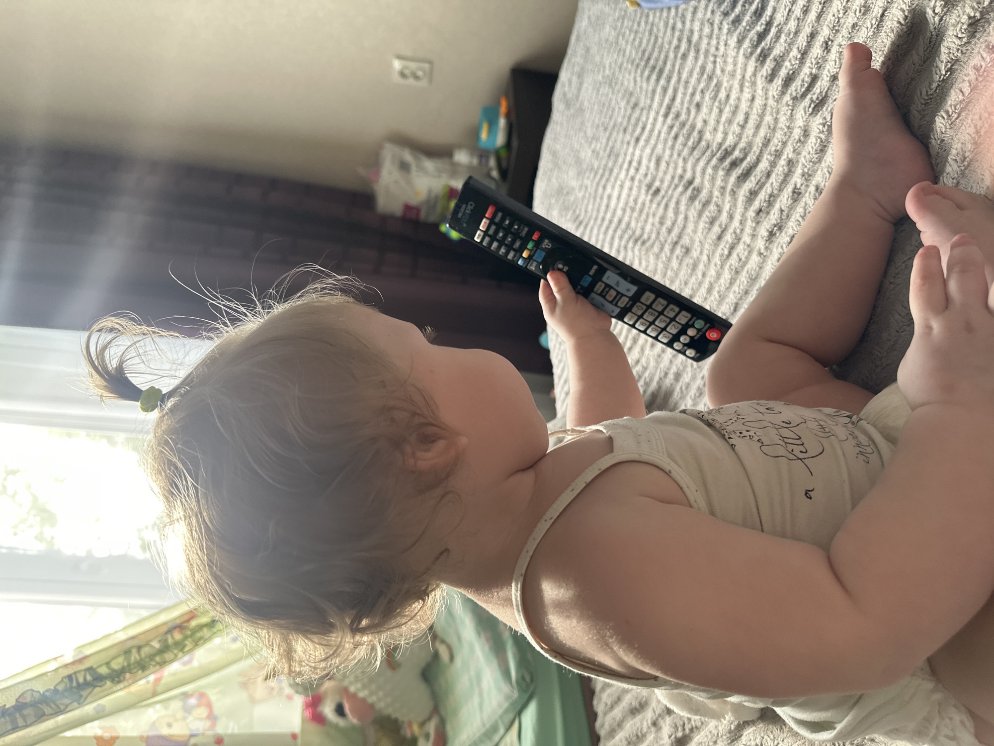 The image shows a toddler, seen from the side, sitting on a bed and holding a television remote control. The toddler has blond hair pulled into a ponytail and is wearing a light-colored tank top. The bed is covered with a gray, textured comforter. In the background, sunlight streams through a window with sheer curtains, illuminating the room. A dark curtain hangs to the right of the window. A bedside table with some items on it is visible in the distance, and a light switch is mounted on the wall. The overall scene appears to be in a domestic setting, likely a bedroom. The image does not contain any specific visual elements that would allow for an accurate determination of its location within the city of Sayanogorsk, Russia.