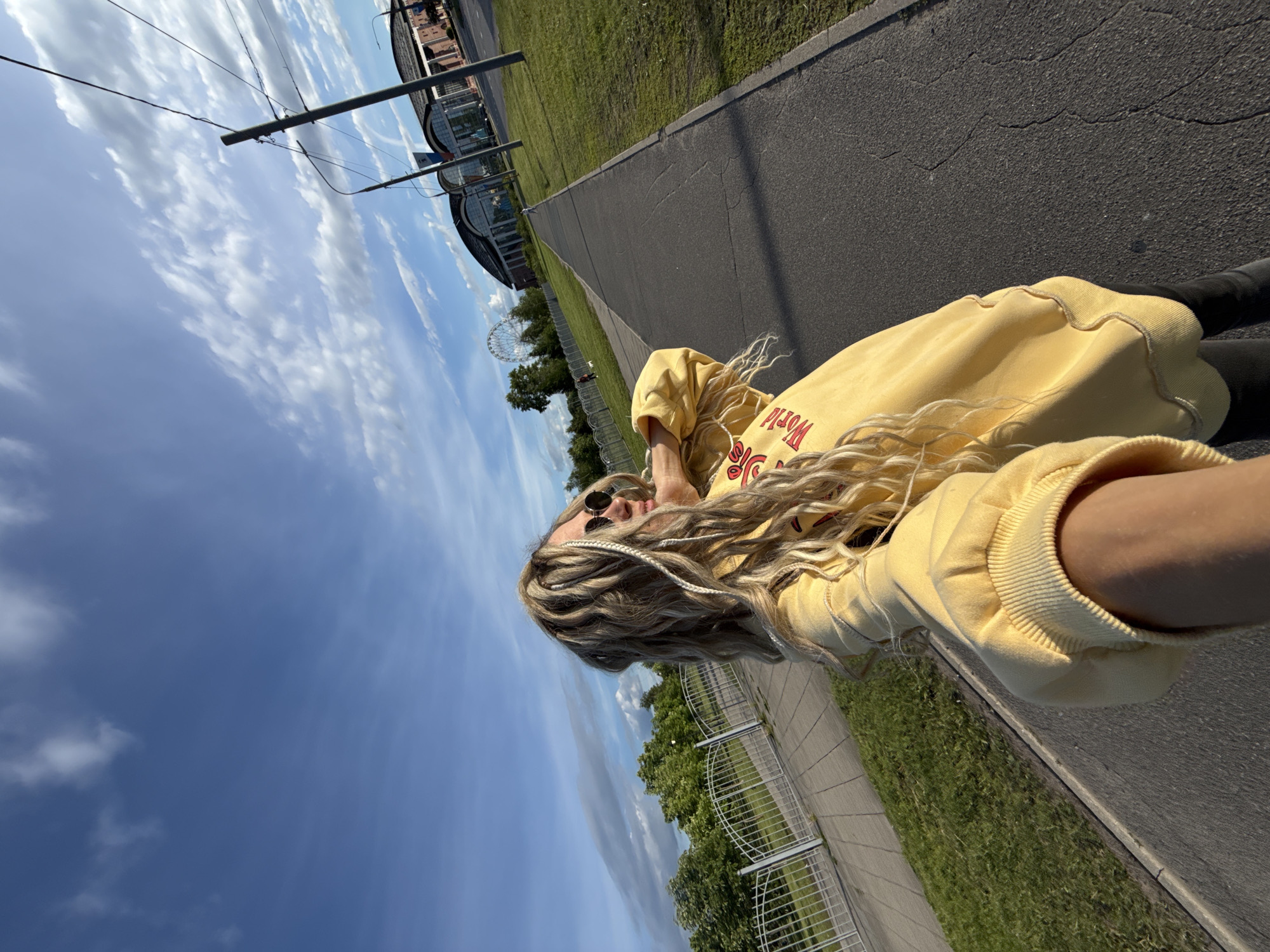 A medium-angle outdoor shot shows a woman in a yellow sweatshirt with red writing. She is wearing sunglasses and has blonde hair with braids. She appears to be taking a selfie, as her arm is outstretched towards the camera.
The environment includes a gray asphalt path, green grass, a metal fence, trees, and buildings in the distance. A Ferris wheel is also visible in the background. Overhead power lines are present. The sky is blue with scattered clouds.
The location is potentially in the city of Шебунино, Russia, given the request.