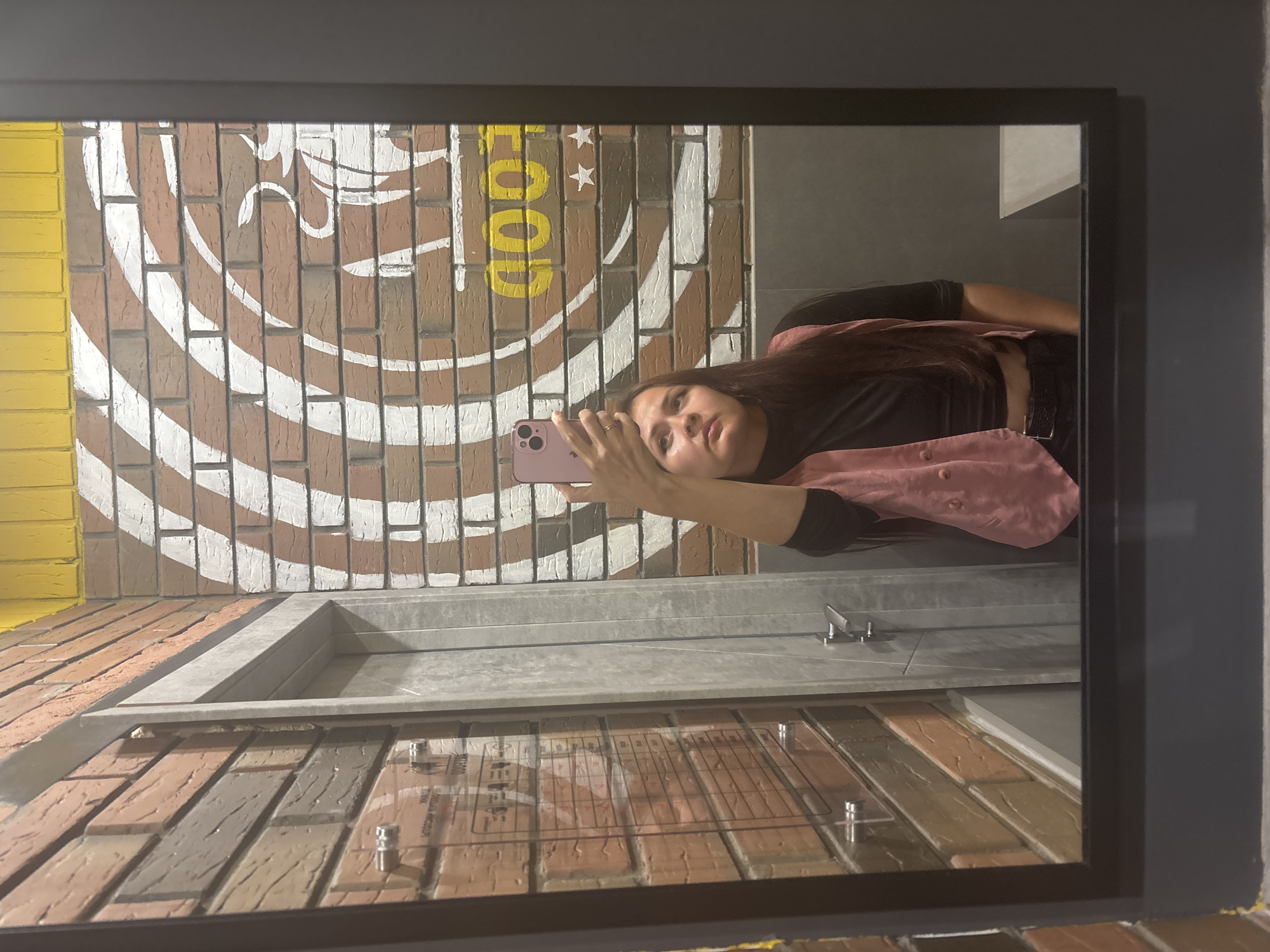 A woman stands in front of a mirror in a restroom. She is taking a selfie with her pink smartphone, which she holds up with her right hand. She is wearing a black long-sleeved crop top, a pink vest, and dark pants with a belt. Her hair is long and brown. The restroom has a brick-patterned wall with white and yellow accents, and a yellow ceiling. A door with a metal handle is visible in the background, as is a sign on the wall. The floor appears to be tiled. The mirror reflects the woman and the interior of the restroom. The location is identified as Naberezhnye Chelny, Russia.