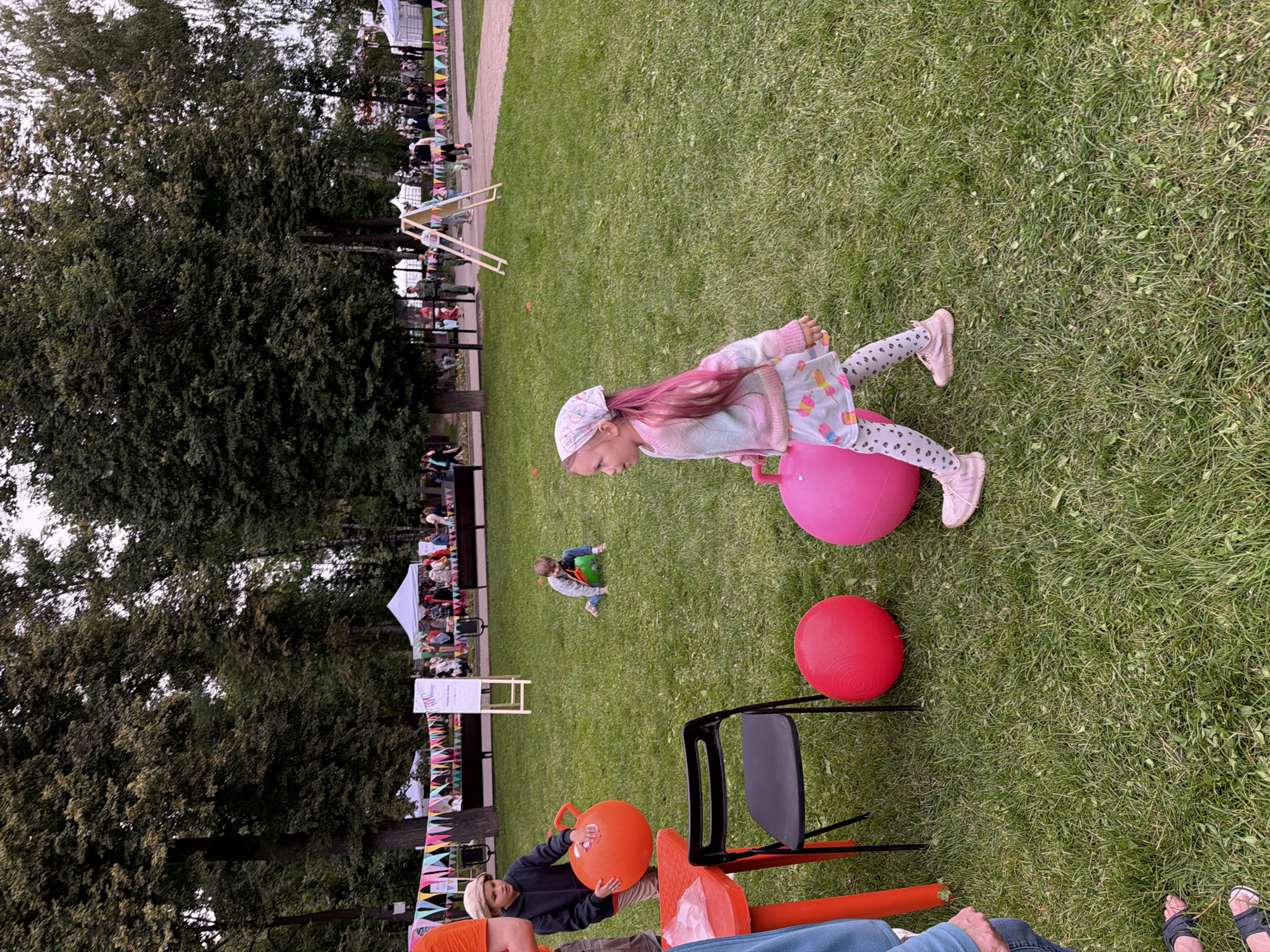 The image depicts an outdoor event likely a festival or fair taking place on a grassy field in Kazan, Russia. In the foreground, a young girl with pink hair is joyfully bouncing on a large pink inflatable ball. She is wearing a light-colored top with colorful patterns and patterned leggings, along with pink shoes. Nearby, another child is also playing on a similar inflatable toy, a green one.
The background shows a gathering of people, some sitting and watching, while others are milling around. There are tents and temporary structures, suggesting a festive atmosphere. A series of colorful triangular flags are strung across the scene, adding to the celebratory feel. Trees provide a backdrop of natural greenery, and the overall lighting suggests it might be late afternoon or early evening. The mood appears relaxed and cheerful, with children actively engaged in play and adults observing. The presence of various inflatable toys indicates a child-friendly activity or entertainment zone.