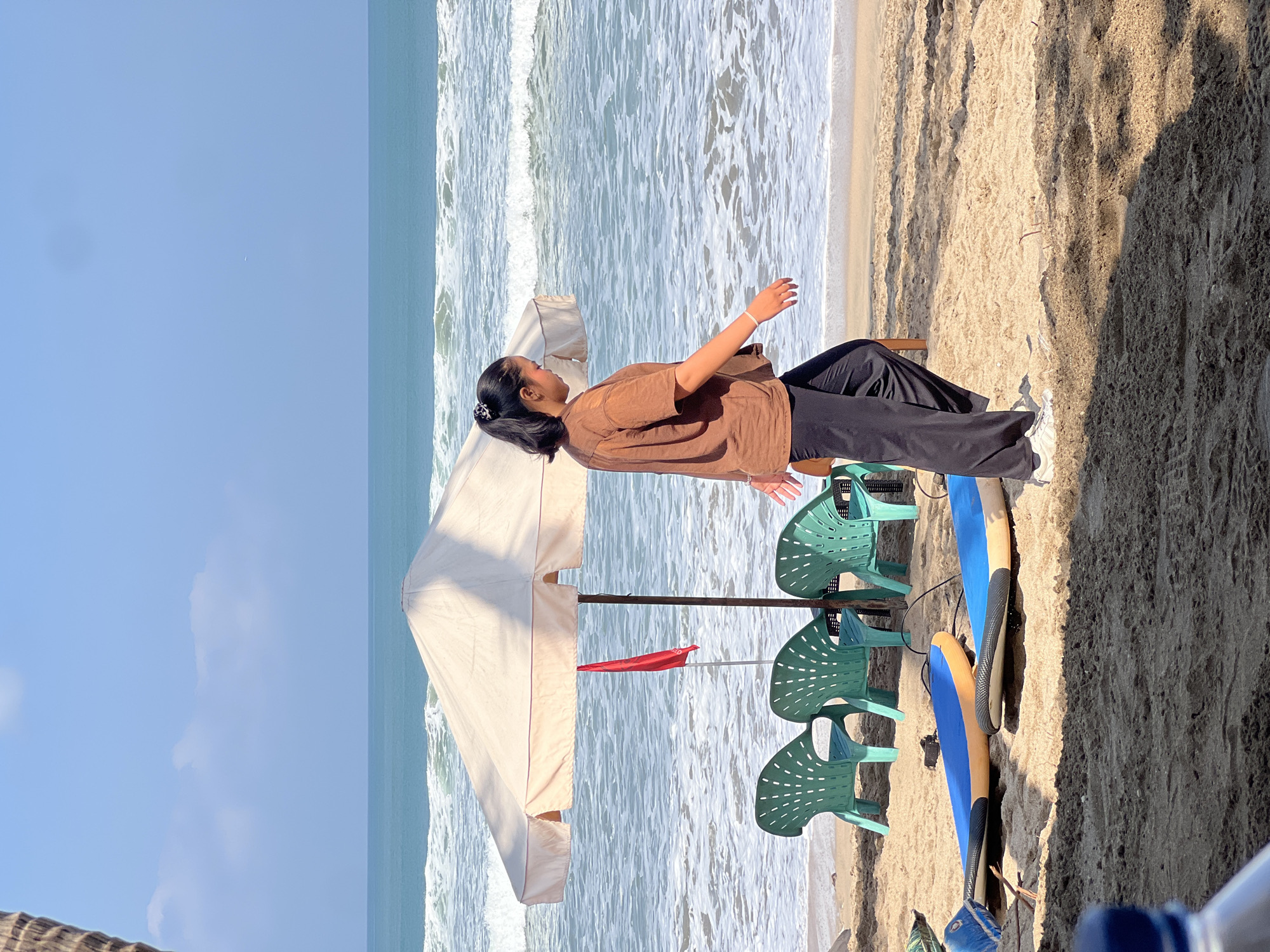 In Kuta, Indonesia, a woman with dark hair pulled into a ponytail walks along a sandy beach. She is wearing a brown t-shirt and black pants. The ocean, with moderate waves, is behind her, and a large, light-colored beach umbrella provides shade over three green plastic chairs and a red flag. Two surfboards lie on the sand in the foreground.