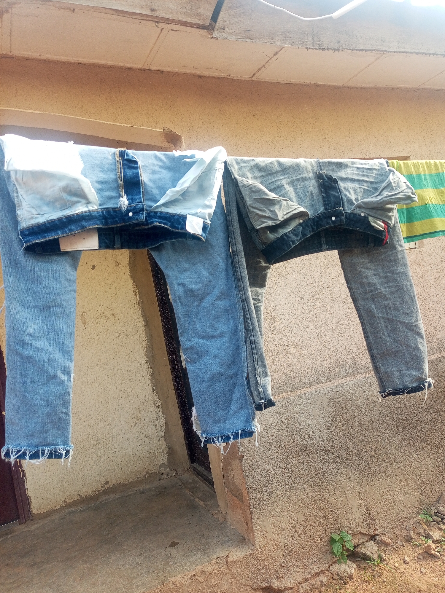 Two pairs of denim jeans are hanging on a clothesline outdoors. The jeans are light blue and appear to be newly washed, as they are draped over the line. The scene is set against a textured, light-colored wall, suggesting an outdoor drying area. The lighting suggests it is daytime, with sunlight casting shadows. In the background, a section of a striped cloth is also visible, possibly another item of clothing or a towel. The overall impression is one of domesticity and routine laundry practices in Jalingo, Nigeria. There is no visible text in the image.