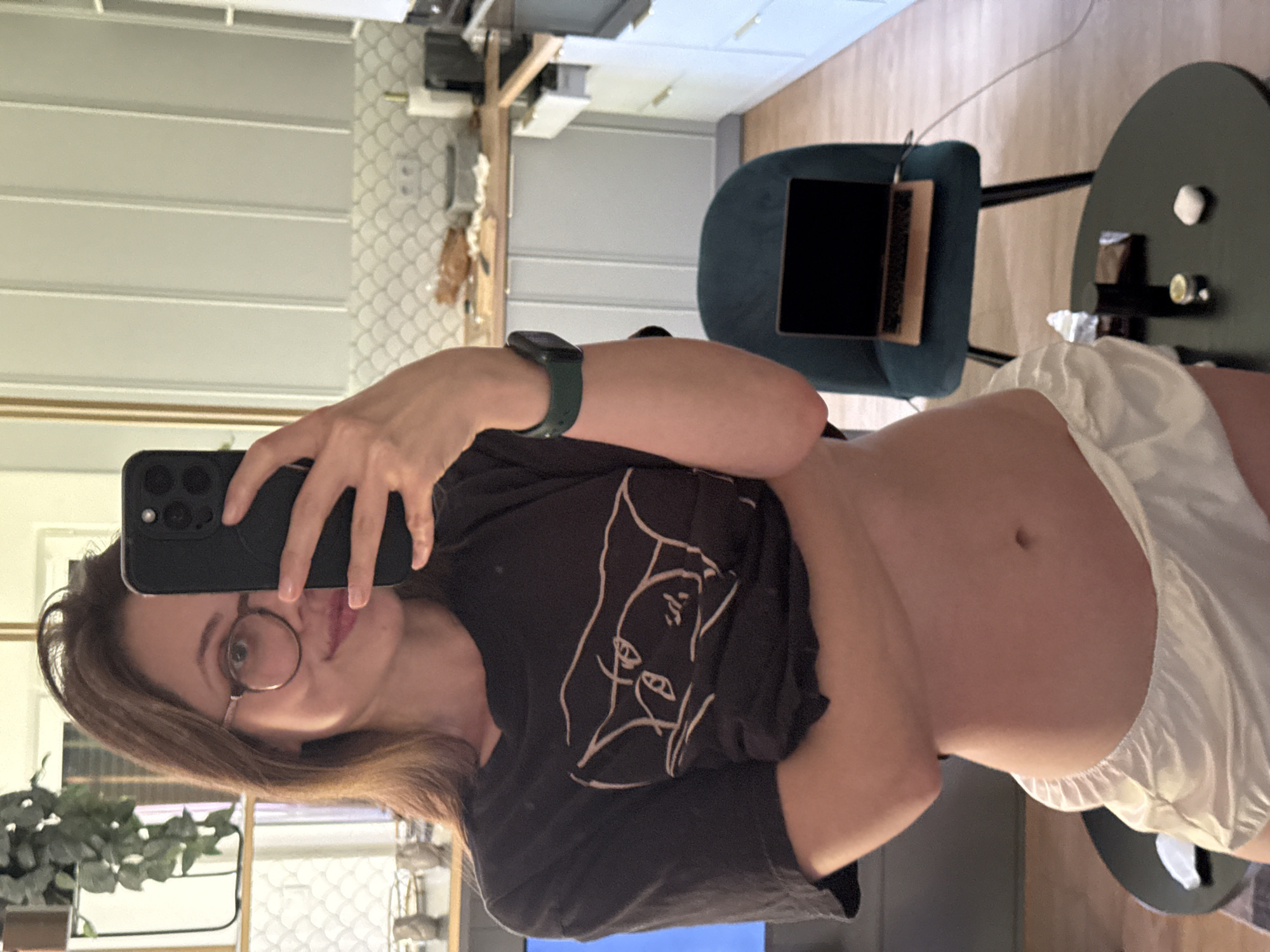 A woman with brown hair and glasses takes a selfie in a kitchen. She is wearing a black t-shirt with a white graphic print and white silk shorts. A dark green smartwatch is on her left wrist. Behind her, a laptop sits open on a dark teal chair. The kitchen has light gray cabinets and a white tiled backsplash. The image appears to be taken indoors, likely in a residential setting. No specific location in Krakow, Poland, is visible.