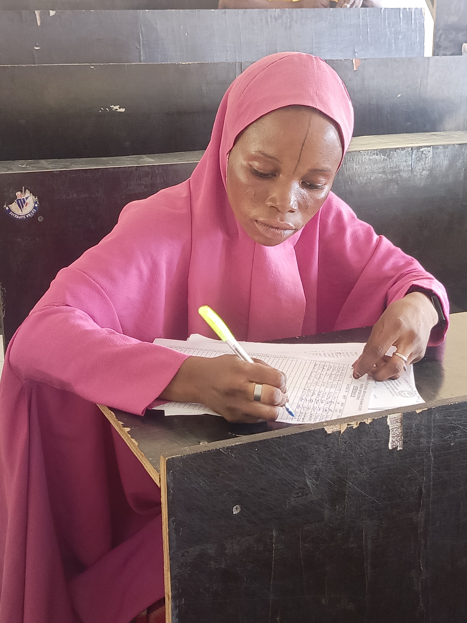 A woman wearing a pink hijab is sitting at a desk and writing on a form with a pen. The form appears to be a written document, perhaps an attendance sheet or a test paper. The desk is worn, with visible damage to the surface. The scene appears to be inside a classroom or office setting in Akwanga, Nigeria, based on the architectural style and form.