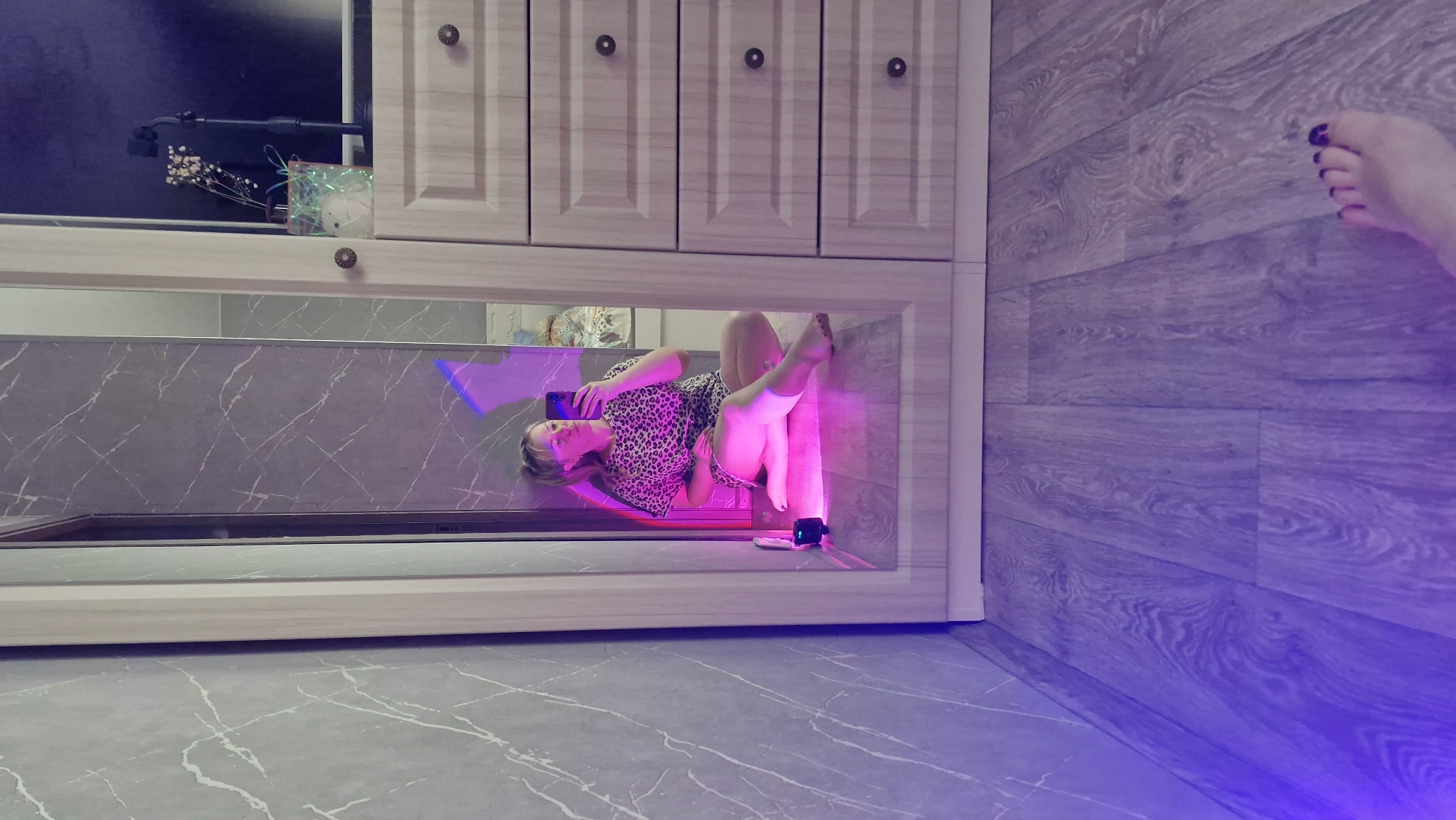 The original image is rotated 90 degrees clockwise, depicting an indoor scene with strong artificial lighting. In the center, a large, rectangular reflective surface, likely a mirror, shows the reflection of a woman. She is wearing a leopard-print dress or top, posing with her legs slightly bent. She holds a smartphone, seemingly capturing a self-portrait in the mirror. A prominent pink and purple light illuminates the reflected scene, casting vibrant hues on the woman and the marble-patterned surface behind her in the reflection.
Above the reflective surface are several light-colored wooden cabinets, each with a dark, round knob. On a shelf above the mirror, decorative elements include a small vase with dried white flowers and a glowing, translucent box. On the surface directly in front of the mirror (within the reflected area), a small black device, possibly a camera or an additional light source, is visible.
To the right side of the original image, which would be an adjacent wall or floor if properly oriented, there is a section of wooden flooring or paneling with a light gray, plank-like pattern. A woman's foot, with dark nail polish on the toes, extends from the top-right corner into this area. The overall setting, characterized by the mirror, specific lighting, and posing, suggests a photoshoot or video recording in an evening indoor environment. No text is visible in the image. The location is Otchor, Russia.