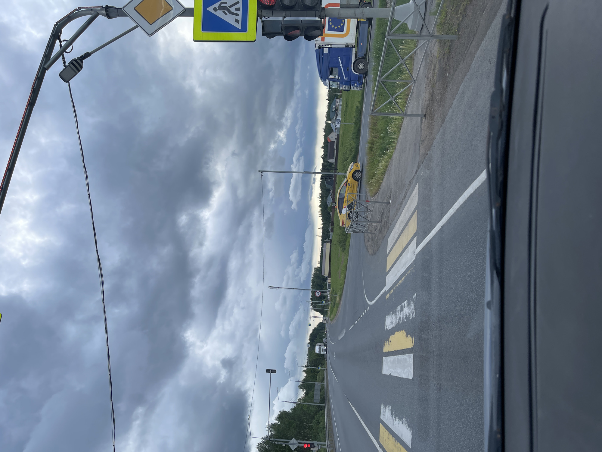 The image, apparently taken from inside a vehicle, shows a view of a road in Novyi Byt, Russia. The road is asphalt and has white and yellow lines marking the lanes. A pedestrian crossing with white and yellow stripes is visible. A yellow taxi cab is parked on the side of the road. Traffic lights and road signs, including a pedestrian crossing sign and a diamond-shaped sign, are present. A blue truck with the EU flag on it is also parked on the side of the road. The sky is overcast with heavy clouds. Trees and other vegetation line the road. The image is rotated counter-clockwise.