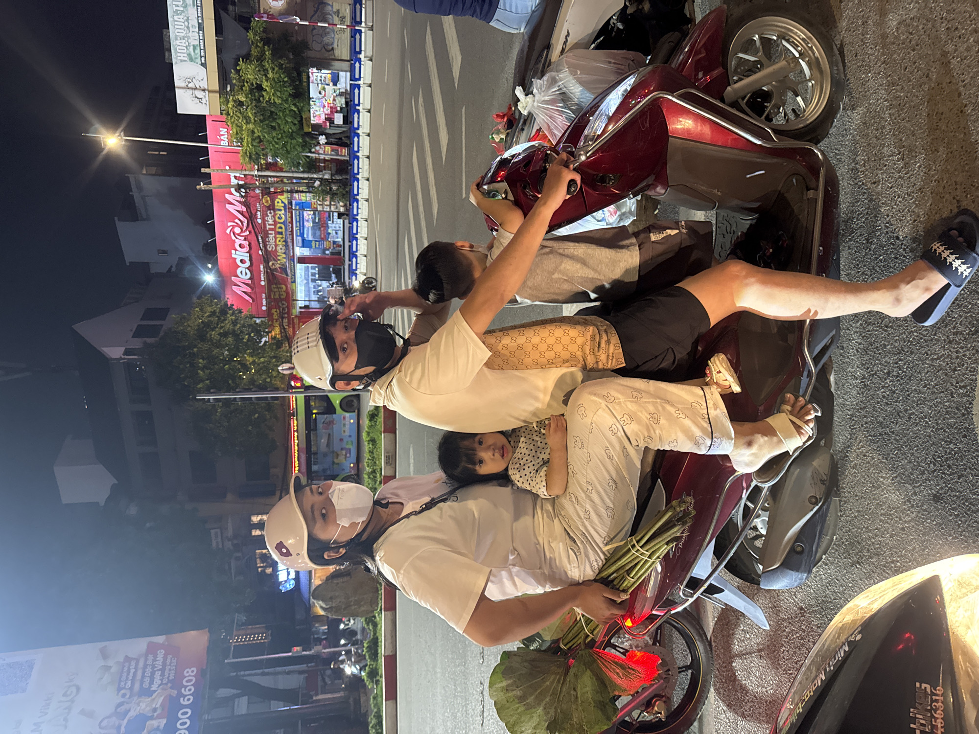 A family of four, two adults and two children, are riding a maroon scooter in Hà Nội, Vietnam. The scooter is in motion on a paved road at night. The adult male in the front is wearing a white t-shirt and black shorts and a beige helmet. The adult female behind him is wearing a white t-shirt and white pants with a bunny pattern and a beige helmet. A young boy is seated behind the adult male, wearing a grey t-shirt and a beige helmet. A young girl is seated in front of the adult female, wearing a polka dot shirt and looking towards the camera. The adult female is holding a large bunch of lotus stems. In the background, there are buildings with illuminated signs, including "MediaMart". The scene appears to be a street in a commercial area of the city.