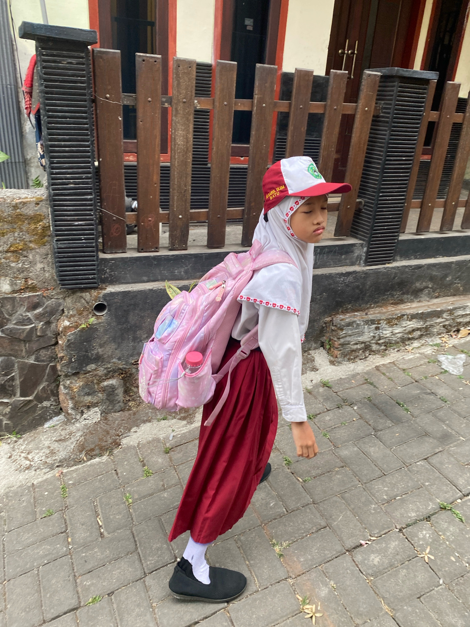 A young girl wearing a white hijab, white blouse with red trim, and a maroon skirt walks on a paved sidewalk. She has on a red and white baseball cap and carries a pink backpack with a water bottle. The girl is walking away from the camera. A wooden fence with metal slats is behind her, and a stone wall is to her left.