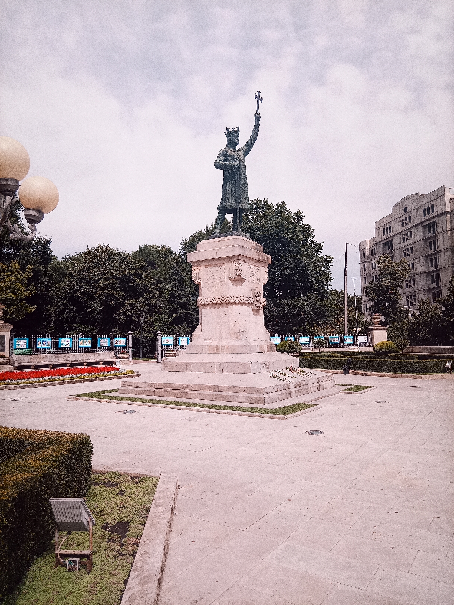 The image shows the monument to Stephen the Great in Chișinău, Moldova. The bronze statue depicts Stephen the Great, a medieval ruler, standing with his right arm raised, holding a cross. He wears a crown and regal attire. The statue is mounted on a large, multi-tiered stone pedestal. In the foreground, there is a paved plaza with some manicured bushes and a street lamp. Beyond the monument, there are trees and a large, unfinished building with multiple windows. The sky is overcast. There are no people visible in the image.