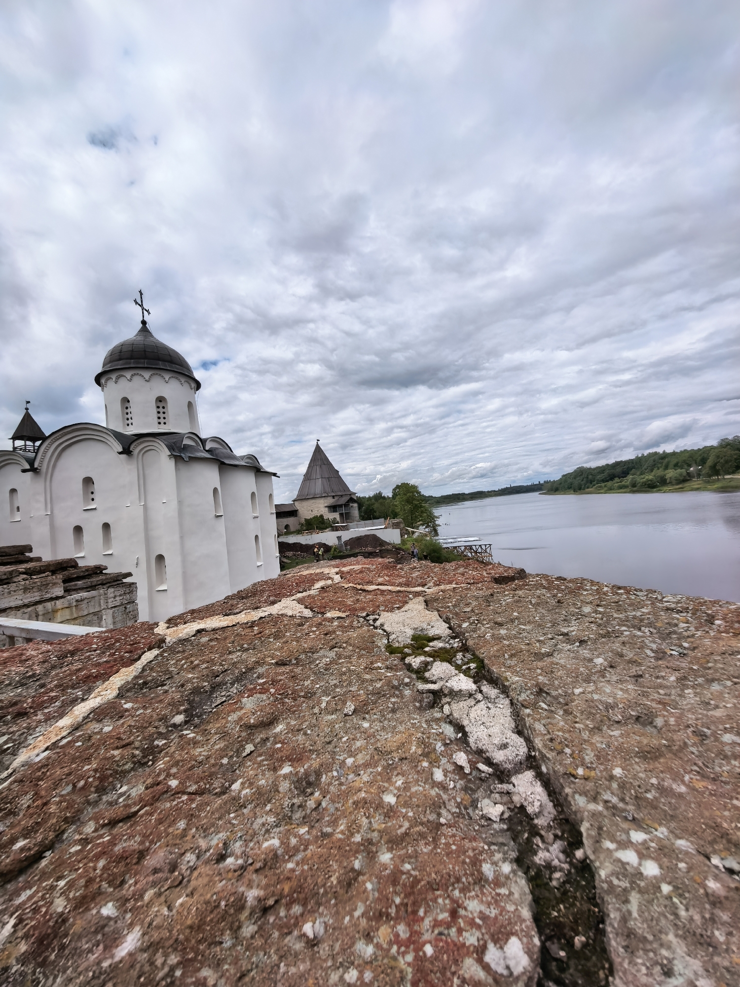 A low-angle, wide-shot photograph captures a view from atop a weathered, reddish-brown stone wall, looking towards an ancient white church with a dark dome and a cross on its peak. The church is situated on a slight elevation, with several arched windows and smaller rectangular openings visible on its facade. To the right of the church, a conical wooden tower stands, part of a larger fortress complex. The scene is dominated by a vast, overcast sky filled with heavy, gray clouds that extend to the horizon. Beyond the fortress, a wide river flows, its surface reflecting the muted sky. The far bank of the river is lined with a dense forest of green trees. The stone wall in the foreground shows signs of age and erosion, with visible cracks and patches of moss. The overall atmosphere is one of historical depth and a somber, cloudy day in Staraya Ladoga, Russia.