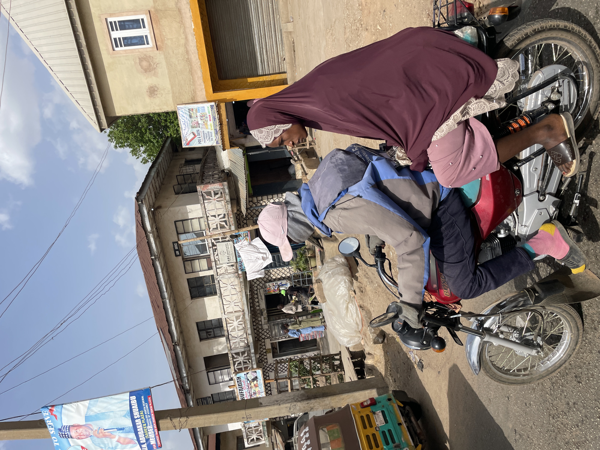 The image captures a vibrant street scene in Jos, Nigeria, under bright daylight with a clear blue sky. Two individuals are seated on a red and silver "BOXER" brand motorbike, which dominates the mid-ground.
The person operating the motorbike, positioned in front, wears a grey and blue jacket, dark trousers, and a light pink cap. They have distinctive yellow and pink socks worn with dark sandals. Behind them, a passenger, presumably a woman, is clad in a long, deep plum-colored abaya or hijab, featuring white lace detailing around the face. She is also wearing dark sandals and appears to be looking forward or slightly downwards.
The background features several multi-story buildings. To the left, a light yellow building with a white-framed window is visible. Below it, an older building with multiple balconies and decorative metal railings showcases clothes hanging to dry. Various shopfronts and general street activity are hinted at between the buildings. Overhead, power lines crisscross the expanse of the bright sky, which has a few scattered clouds.
Notable details include a large blue and white political banner hanging on the left side of the frame, promoting "ALH. ABUBAKAR SHUAIBU" for "MEMBER, NORTH FEDERAL ASSEMBLY," and mentioning "SADEEQ PLAZA" and "SADIQ GOJE (SARKIN YAKIN PLAZA)". A shop sign advertises "SALADIMA enterprises" offering "POS & DATA WITHDRAWAL TRANSFER PAY BILLS." Another sign partially reads "RUFAIDAR Dadi," and text "LAW OFFICE ABC ADVOCATES ABURI" is visible on a building facade. The overall scene depicts a typical moment of daily life and commerce in an urban Nigerian setting.