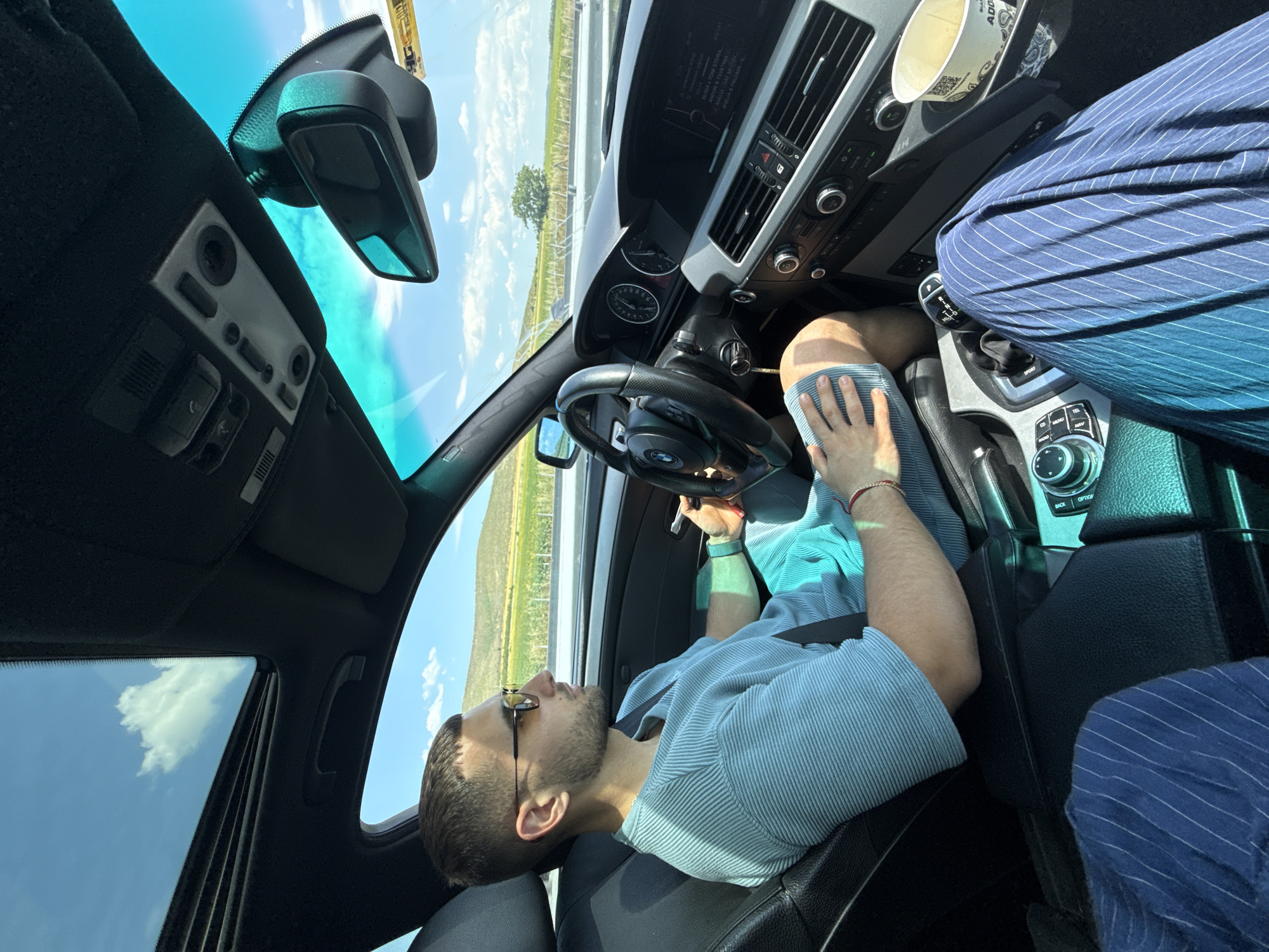 The image depicts the interior of a car on a sunny day. A man with short dark hair, wearing sunglasses, a light blue striped shirt, and shorts is driving. He has a seatbelt fastened. The car's dashboard, steering wheel, and gear shift are visible. Outside the car, a scenic landscape of rolling hills and fields under a bright blue sky with scattered clouds can be seen. The location is identified as Polsko Padarevo, Bulgaria.