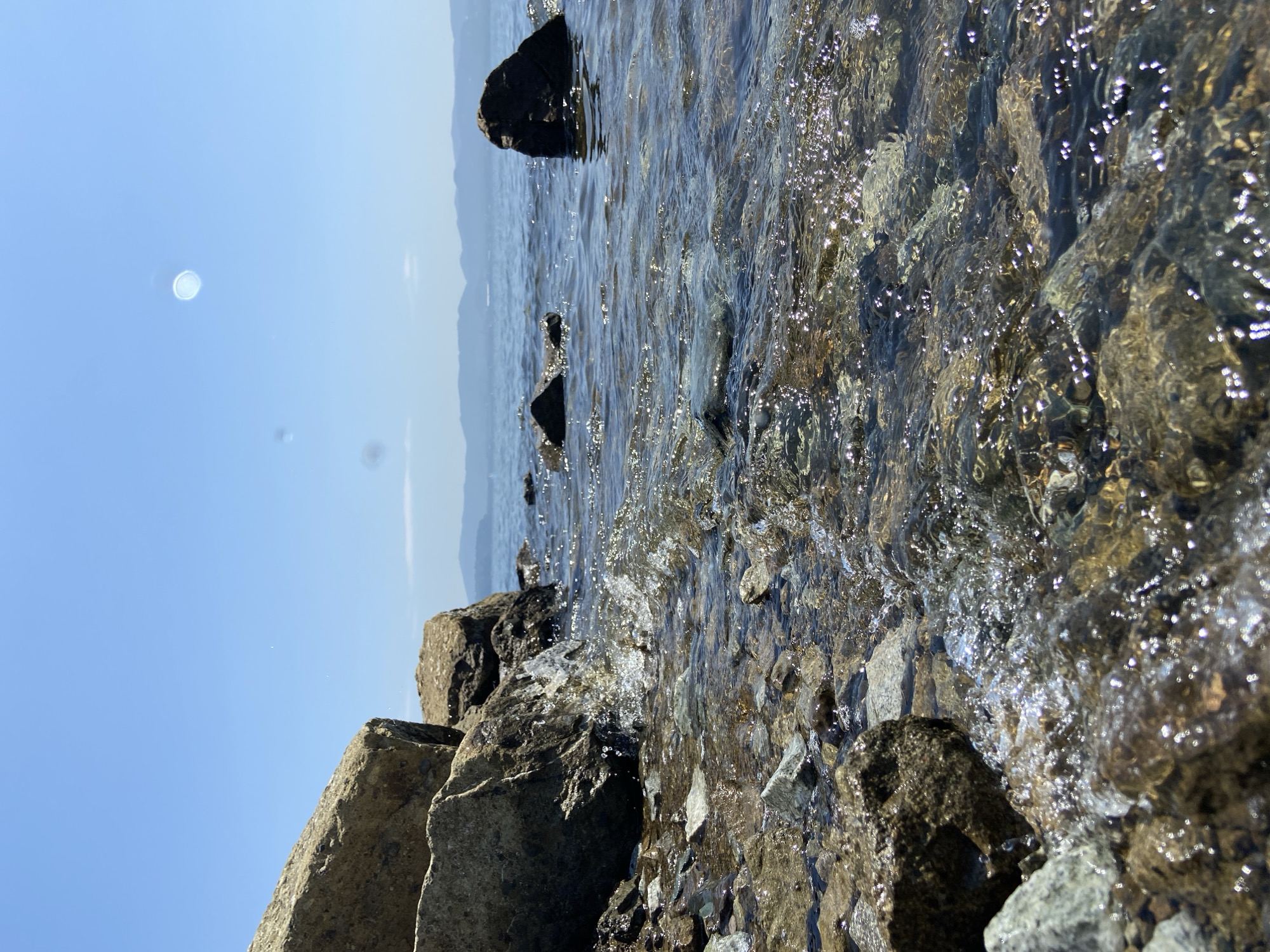 The image depicts a close-up view of a rocky shoreline on a clear, sunny day. Large, weathered rocks are positioned on the left side of the frame, partially submerged in shallow, clear water. The water's surface is rippled, with sunlight glinting off the small waves and illuminating the multicolored pebbles and rocks beneath the surface. In the background, the calm sea stretches to the horizon, meeting a distant, hazy mountain range under a bright blue sky. There are no people visible in the image. The overall scene is natural and serene, characteristic of the coastal areas surrounding Vladivostok, Russia, such as the shores of the Peter the Great Gulf.
