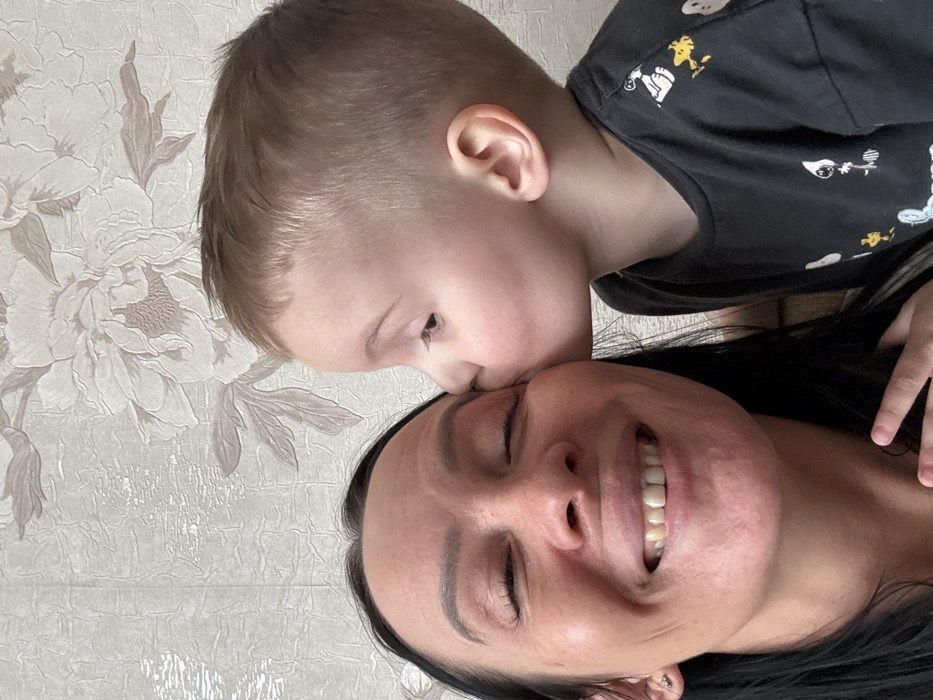 An indoor close-up photograph, situated in the city of Pavlovsky, Russia, presents two individuals. The left portion of the frame features a female with dark hair and light skin, depicted from the chest up. She is smiling broadly with closed eyes, revealing her teeth. A small stud earring is present in her visible left earlobe. Fingers from her right hand are partially visible at the bottom edge. To her right, a young male child with short, light brown hair and light skin is shown in profile, leaning towards the female and making contact with her right cheek/temple area, consistent with a kiss. The child is wearing a dark t-shirt patterned with multiple light-colored cartoon characters identified as figures from the Peanuts comic strip, including Snoopy and Woodstock. The background comprises a wall covered in light-toned, textured wallpaper featuring a repeating pattern of large, muted floral designs.