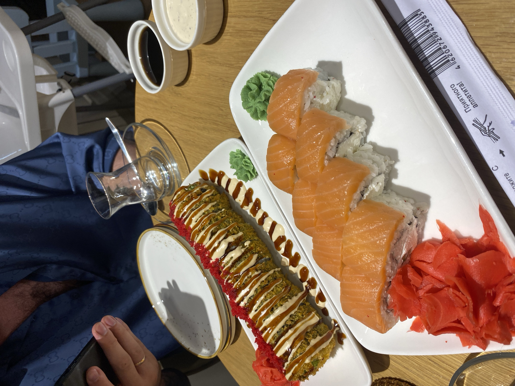 The image shows two plates of sushi on a wooden table. One plate contains a long, cylindrical sushi roll topped with red roe and drizzled with sauce, accompanied by pickled ginger and wasabi. The other plate holds several pieces of salmon nigiri with a dollop of wasabi. To the right, there are two small white bowls, one containing soy sauce and the other a creamy dipping sauce. In the background, a person wearing a blue patterned shirt is visible, holding a smartphone. A glass teacup and a child's high chair are also present. The scene suggests a dining experience at a restaurant in Makhachkala, Russia.