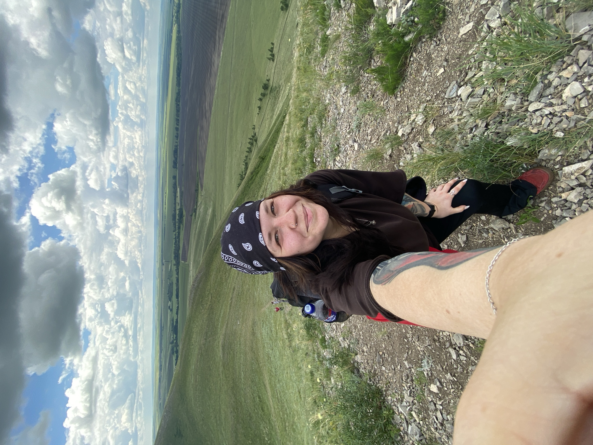 A young woman is taking a selfie on a rocky hillside overlooking a vast, agricultural landscape. She is wearing a dark brown long-sleeved shirt, black pants, and a black bandana with a white paisley pattern. A backpack is visible on her back, with a blue water bottle attached. She has tattoos on her arm, and is wearing a silver bracelet. In the background, several other hikers are visible walking down the hill, with a wide expanse of green fields and cultivated land stretching to the horizon under a cloudy sky. The location is the vicinity of the village of Абзаново, Russia.