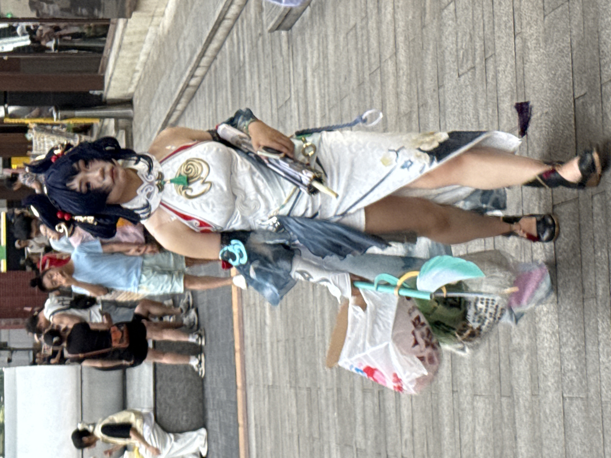 A young East Asian woman, dressed in a white and blue anime-inspired outfit with intricate gold and red detailing, walks along a paved sidewalk in Yuzhong District, China. Her dark hair is styled in an elaborate updo with red accents. She carries a small, folded umbrella in her right hand and is pulling a large, partially filled plastic bag with her left arm. The bag contains various items, including what appears to be a pink stuffed animal. She is wearing black, wedge-style sandals with ankle straps. In the background, other pedestrians are visible, some sitting on a low wall, others walking. The environment suggests a public urban area, possibly a market or pedestrian street, with storefronts and brick structures in the distance.