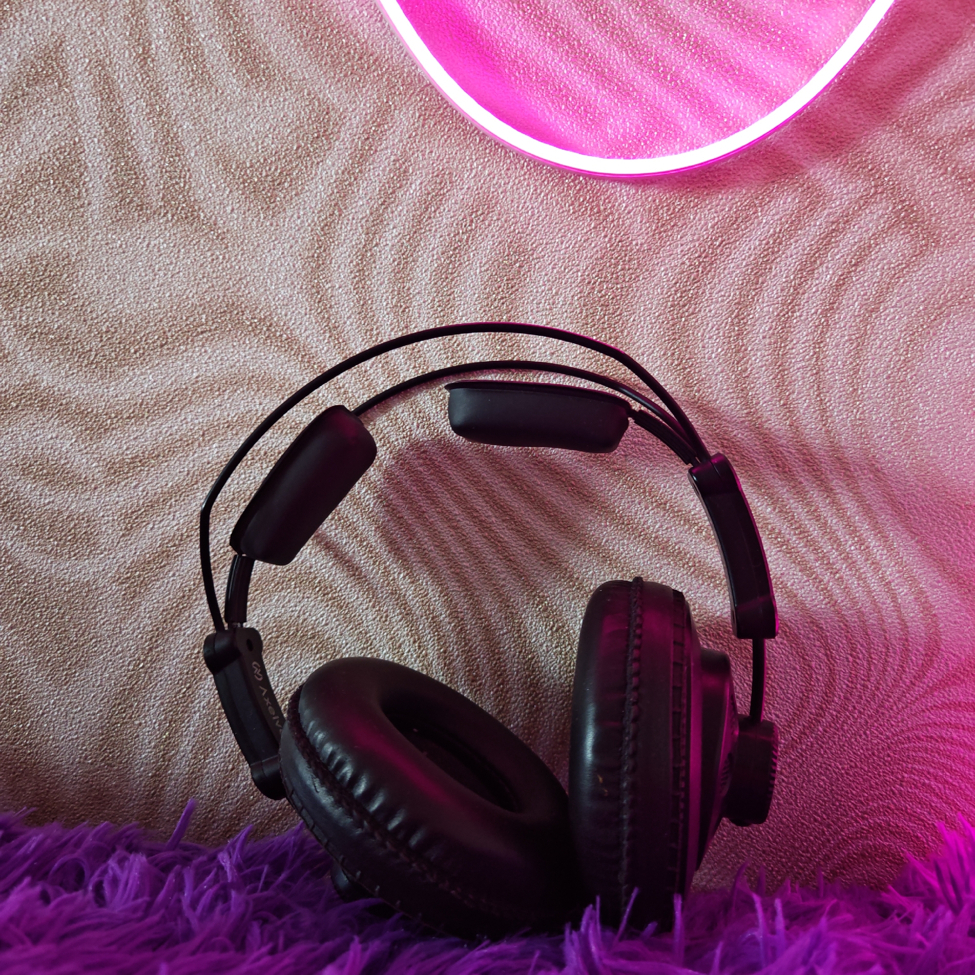A pair of black headphones is placed on a purple shag carpet in front of a tan textured wall with a pink neon light partially visible at the top. The location is Rostov, Russia.