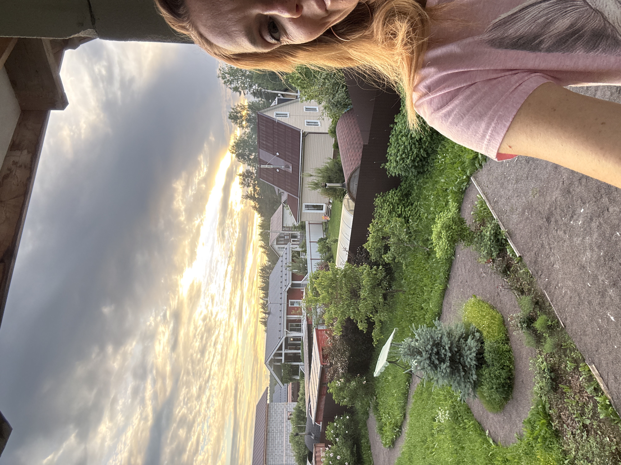 The image depicts a young woman with blonde hair, wearing a pink t-shirt, taking a selfie from an elevated position. She is looking directly at the camera with a slight smile. The background reveals a suburban residential area in the city of Steklyannyi, Russia, characterized by several houses with pitched roofs, some painted red and others beige. The properties are surrounded by green lawns, gardens with various plants, and fences. A small evergreen tree is prominently featured in the foreground, positioned on a curved garden path. The sky above is cloudy with patches of sunlight breaking through, suggesting either dawn or dusk. The overall scene conveys a peaceful, domestic setting.