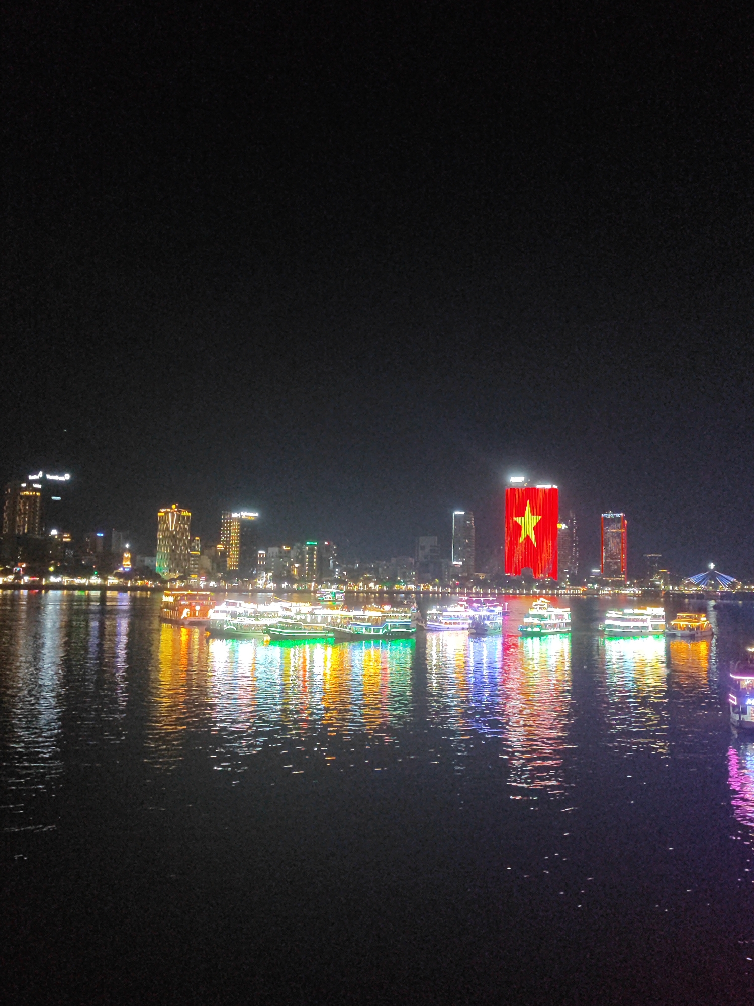 The image shows the city of Đà Nẵng, Vietnam at night. The skyline is illuminated, with several buildings lit up in bright colors, including one tall building displaying the Vietnamese flag in red and yellow. Numerous boats, also brightly lit, are on the river. The water reflects the lights of the city and boats. The sky is dark.