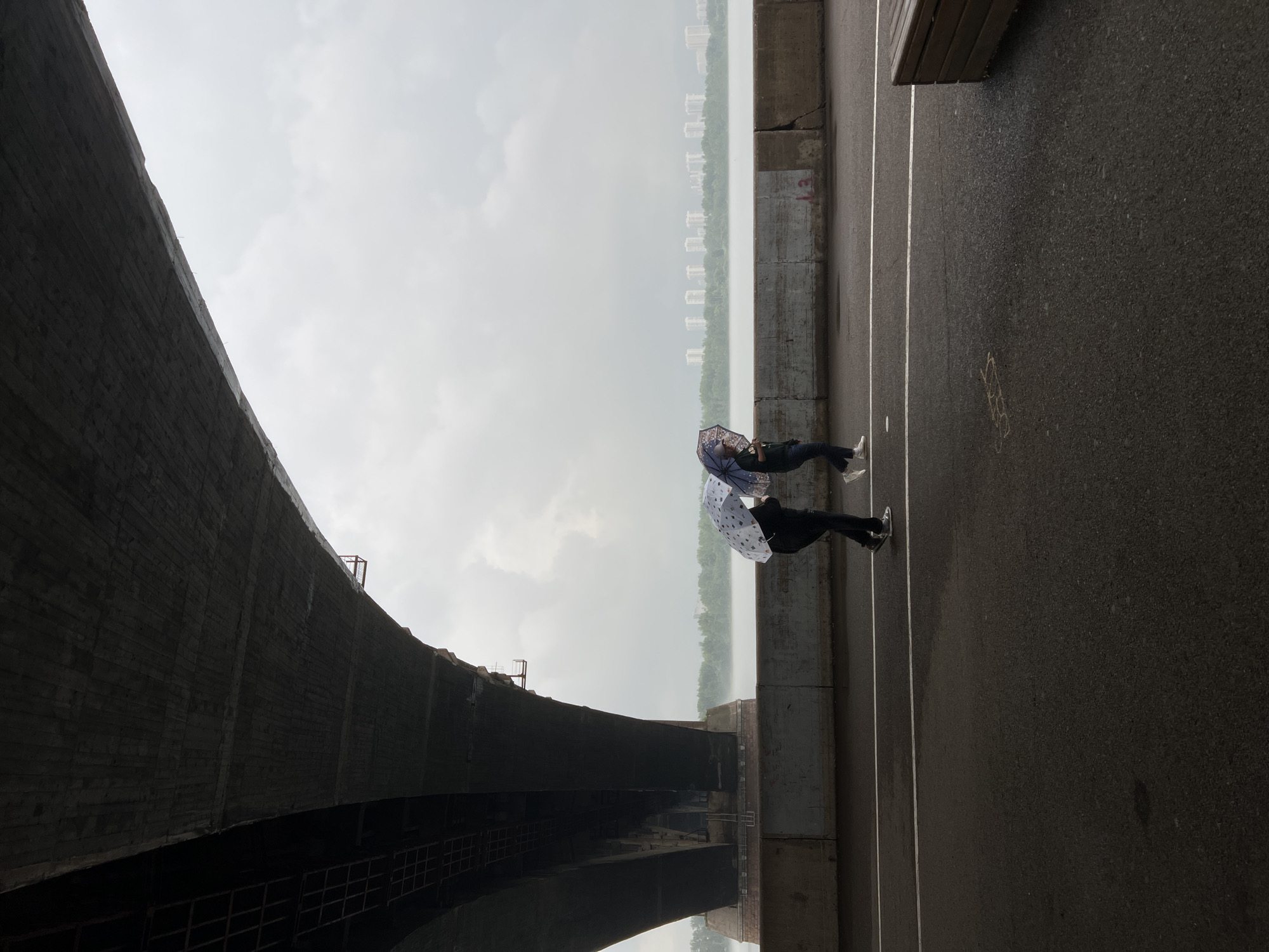 Two individuals are walking under a large concrete bridge in Krasnoyarsk, Russia, on a cloudy day. Both are holding umbrellas, suggesting it is raining or has recently rained. The person on the left is holding a white umbrella with a pattern of dogs. The person on the right is holding a solid dark umbrella. They are walking on a paved pathway that runs parallel to a concrete barrier. Beyond the barrier is a body of water, likely the Yenisei River, with a treeline and a distant cityscape featuring numerous white buildings visible on the horizon. The underside of the bridge is constructed of concrete and features visible structural supports. The foreground is a dark asphalt surface with white lane markings. A wooden bench is visible on the right side of the image.