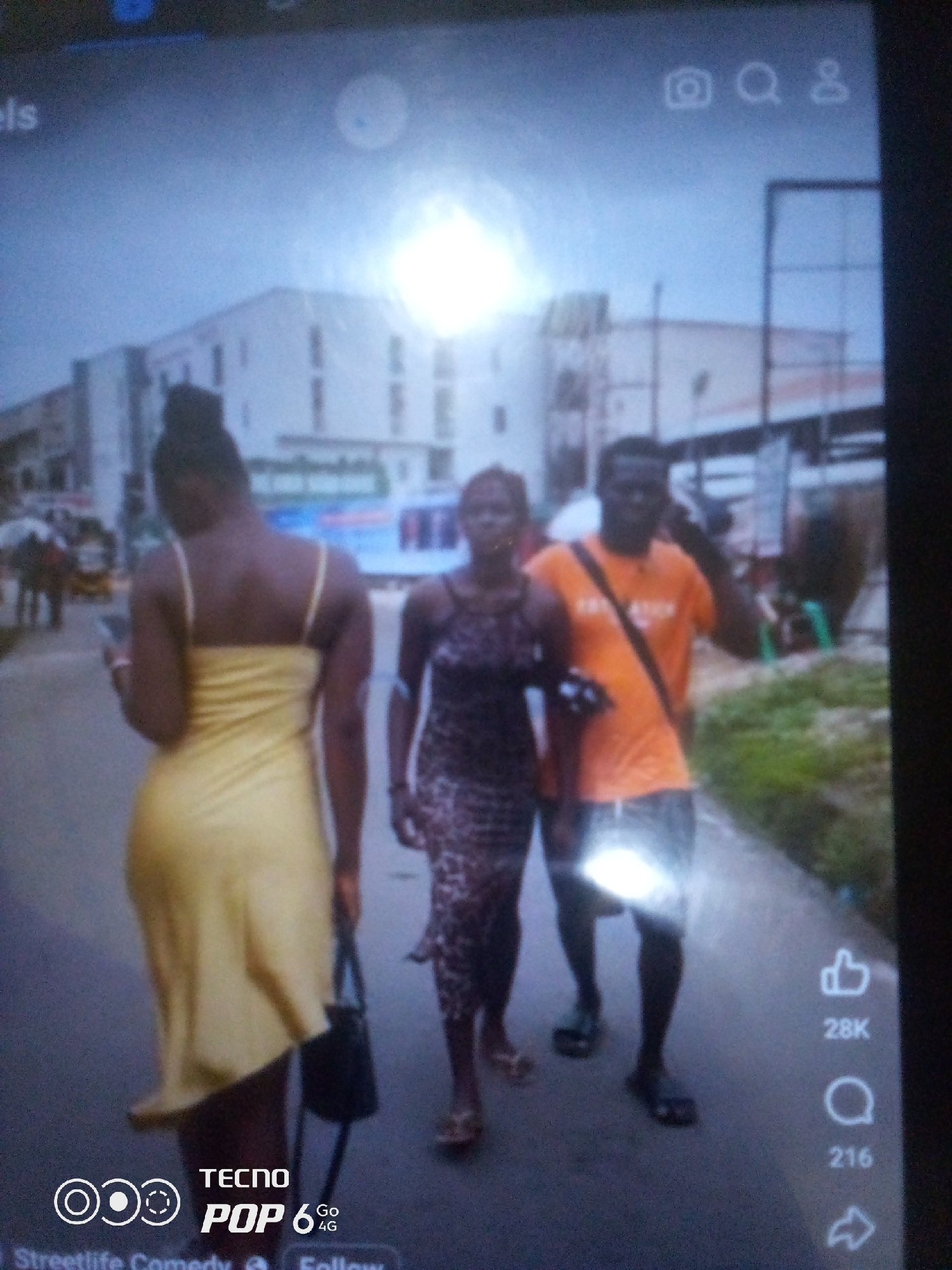 In an urban outdoor setting, possibly Howayi, Nigeria, three individuals are captured walking on a paved pathway. The individual on the left, facing away from the camera, wears a light yellow slip dress and carries a dark handbag. To their right, another individual, facing slightly towards the camera, is dressed in a leopard-print dress and is also walking. The third individual, to the right of the woman in the leopard-print dress, is a man wearing an orange t-shirt with black lettering and dark shorts, with a dark strap across his chest. He appears to be holding a phone to his ear. In the background, a multi-story building with a blue and white facade is visible, along with other structures and some greenery. The lighting suggests it is daytime, with some bright light sources creating glare on the ground and on the subjects. Social media icons, including a camera, a magnifying glass, a user profile icon, a thumbs-up with a count of "28K", a comment icon with a count of "216", and a share icon, are overlaid on the right side of the image, along with "TECNO POP 6 Go 4G" branding at the bottom left.