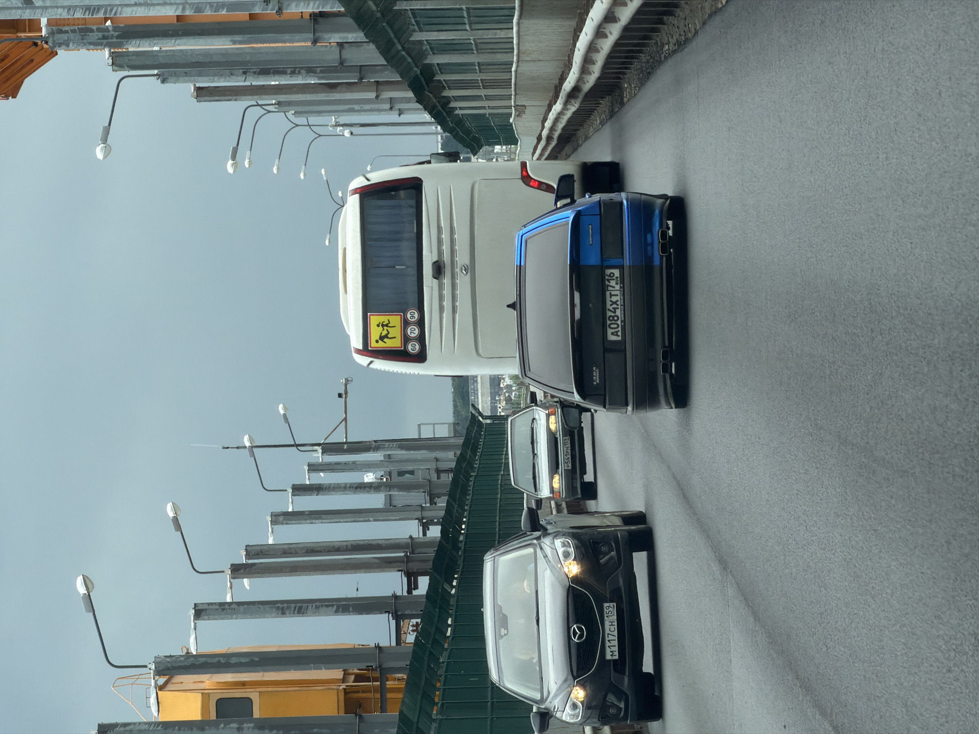 The image depicts a multi-lane asphalt roadway on the Votkinsk Hydroelectric Power Station Dam in Chaikovsky, Perm Krai, Russia, under an overcast sky. Traffic flows away from the viewer.
In the left lane, a dark grey Mazda CX-5 SUV with its headlights on, bearing Russian license plate M117CH 159, is prominently positioned. Two occupants are visible within the vehicle. Further back in the same lane is a silver sedan, likely an older Lada model, with Russian license plate P541PK 159.
In the adjacent right lane, a modified black and blue Lada Samara sedan with dual exhaust pipes and Russian license plate A084XT 716 is visible. Immediately behind this car, a white tour bus or school bus is present, featuring a yellow sign with two stick figures (children) and circular speed limit decals for 60, 70, and 90 km/h on its rear. Behind the bus, another dark vehicle's rear bumper is partially visible.
The roadway is flanked by substantial infrastructure. On the left, a large yellow industrial structure, possibly part of the dam's machinery, stands alongside a series of grey concrete or metal pillars supporting streetlights. On the right, a green mesh fence extends atop a concrete barrier, followed by a parallel row of similar pillars with streetlights. A metal guardrail borders the far right edge of the road. The sky above is a uniform light grey.