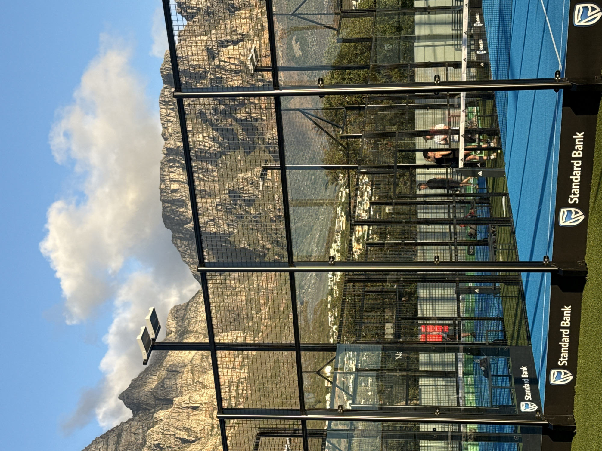 Here is a detailed description of the image:
The image depicts a padel tennis court in Cape Town, South Africa, with Table Mountain prominently in the background. The court features a blue surface and black metal framing, including wire mesh and glass panels. The branding of "Standard Bank" is displayed on the court's structure.
People are playing padel tennis on the court, with several individuals visible in various active poses. The environment includes green grass surrounding the court and buildings in the distance. The sky is blue with scattered clouds, highlighting the natural landscape. A light fixture is also visible to the left of the frame.