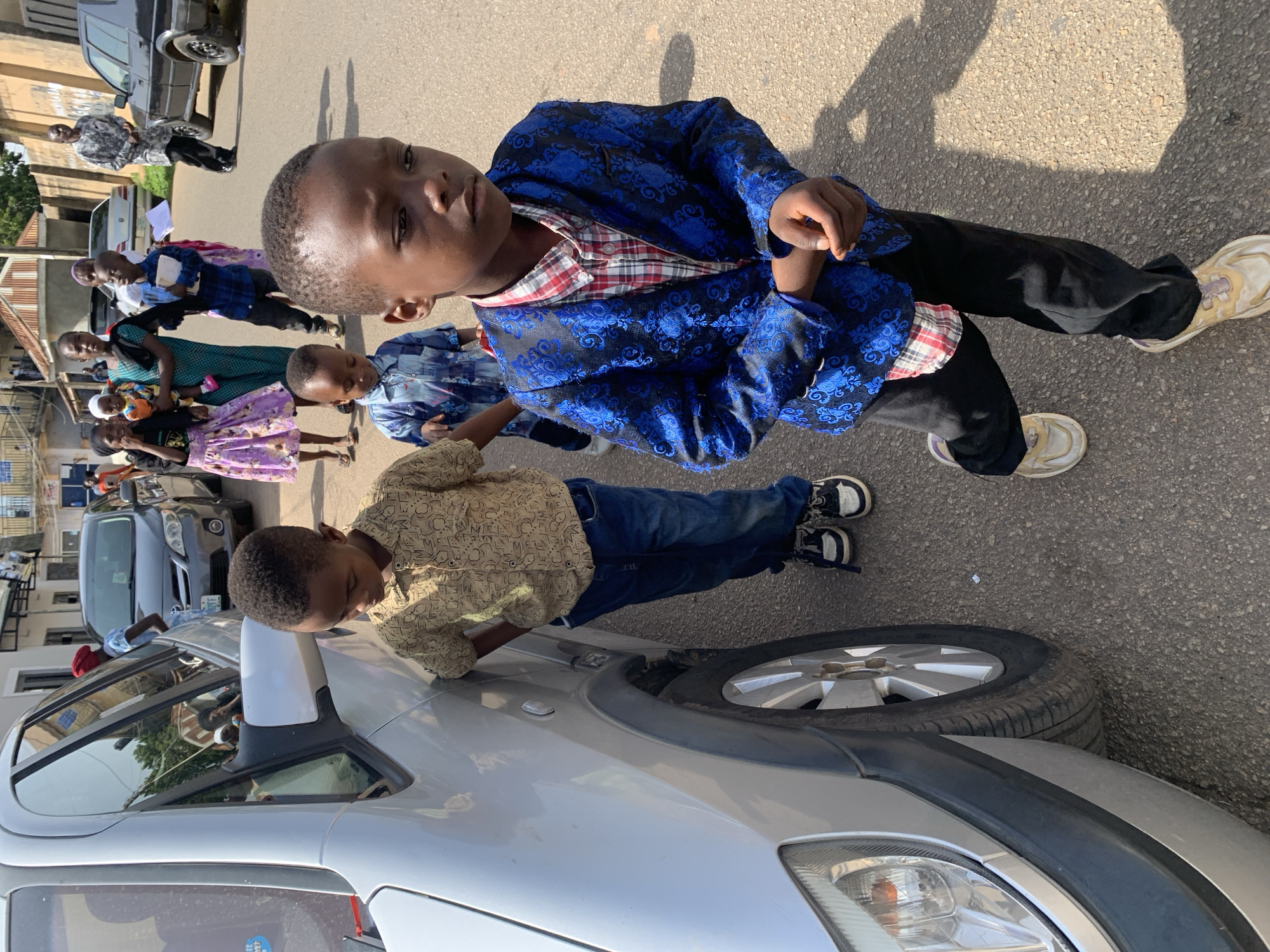In an outdoor, daytime scene in Jos, Nigeria, a group of children and adults are walking down a paved street. In the foreground, a young boy wearing a blue, intricately patterned jacket over a plaid shirt and black pants is the primary subject. His arms are crossed, and he is looking downwards. To his left, another young boy in a patterned blue shirt and jeans stands near a silver car. Behind them, other children and adults are visible, some walking and some standing. A woman in a green dress holds a baby. Further in the background, several cars are parked or driving on the street, and buildings line the roadside. The environment appears to be an urban setting with a mix of residential and commercial structures. The lighting suggests a sunny day, casting distinct shadows on the ground.