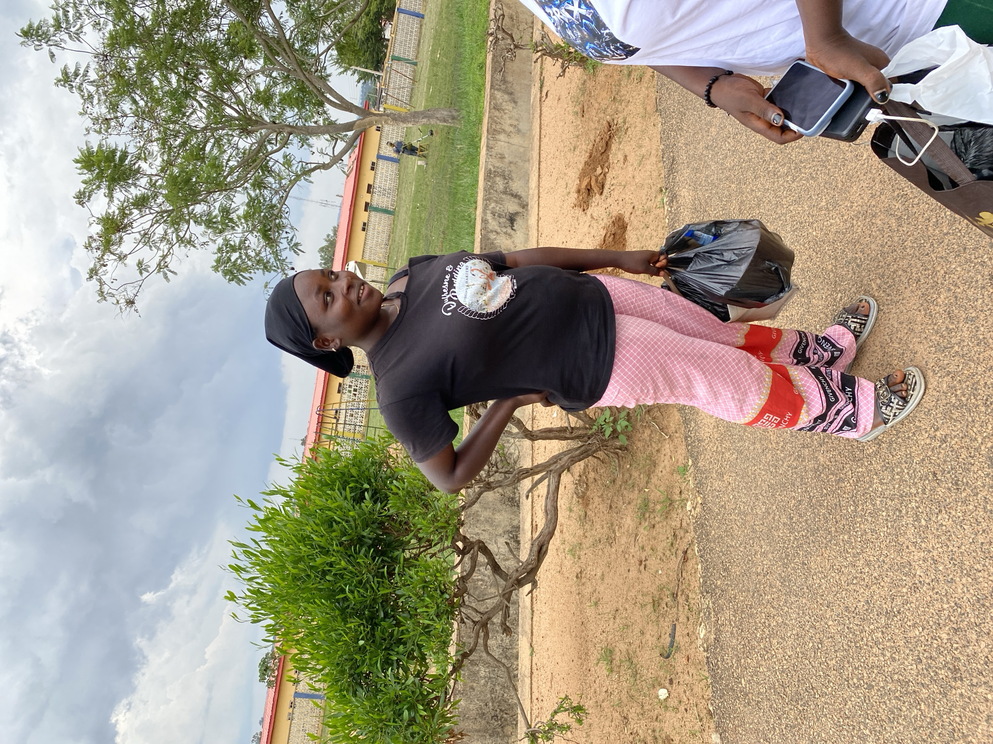 In Vom, Nigeria, a young woman stands outdoors on a paved walkway. She is wearing a black t-shirt and pink checkered pants with red and black accents. She has a black headscarf on and is holding a black plastic bag in her right hand. Her left hand is on her hip. To her right, another person, partially visible, is wearing a white t-shirt and holding two smartphones and a shopping bag. In the background, there is a low building with a red roof, a small tree, and a larger tree. The ground is covered in dirt and sparse grass. The sky is cloudy.