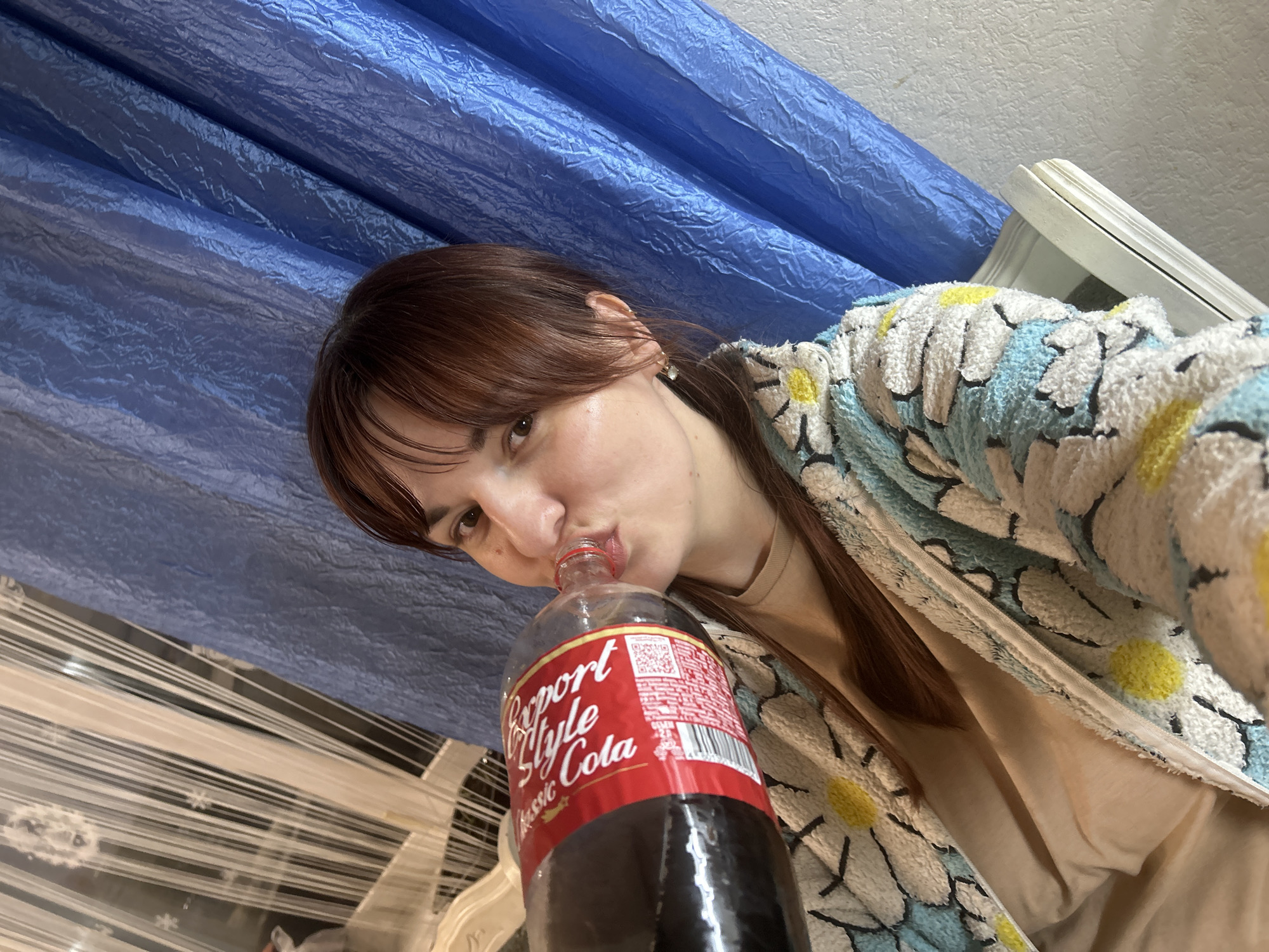 A woman with brown hair is taking a selfie while drinking from a "Classic Export Style Cola" bottle. She is wearing a daisy-patterned robe over a beige shirt. The background includes blue drapery and what appears to be blinds. There's no indication of the location being in Фоки, Russia.