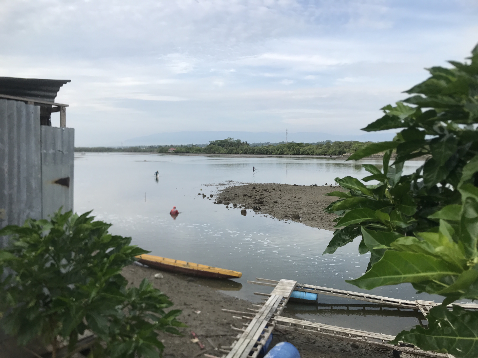 This image depicts a waterfront scene in Lhokseumawe, Indonesia. In the foreground on the left, a weathered structure with a corrugated metal roof and siding is partially visible. In the middle ground, a calm body of water stretches towards a tree-lined horizon with distant hills. A yellow canoe rests on the muddy shore in the lower left of the water. A simple wooden pier with blue barrels beneath it juts out into the water from the right side of the frame. Two figures are visible in the water: one near a red buoy further out in the water, and another in the distance on the right, appearing to be working or fishing. The sky is overcast with scattered clouds. Lush green foliage occupies the right edge of the frame, partially obscuring the view. The scene suggests a coastal or riverine environment, likely a fishing or aquaculture area within the city of Lhokseumawe.