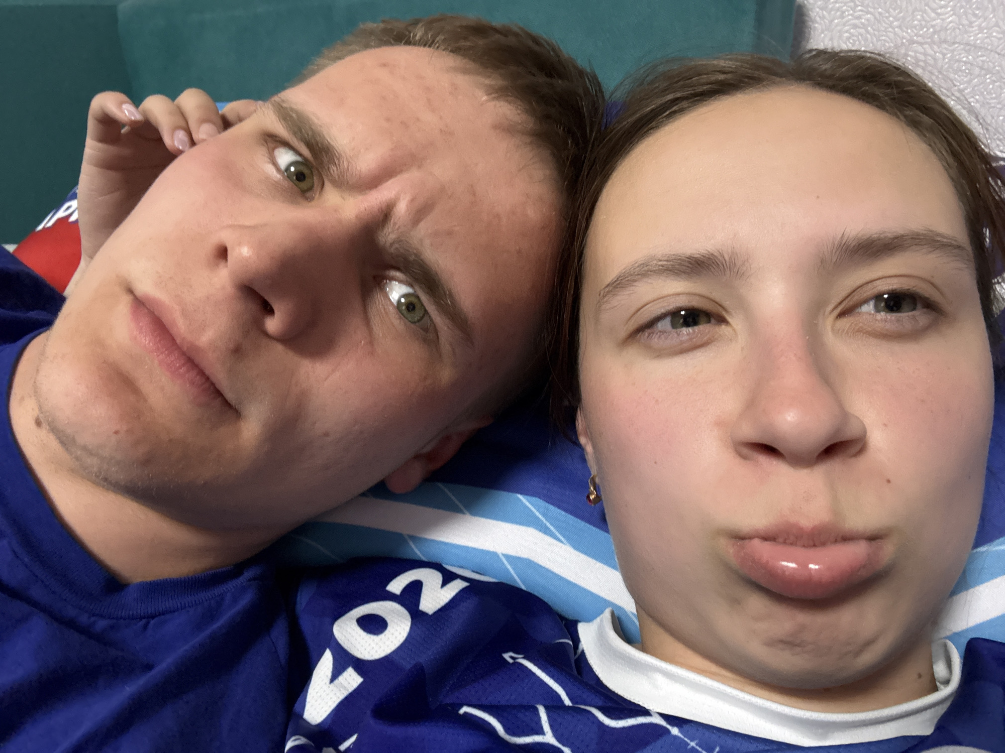 A selfie captures a man and a woman indoors, likely in a home setting in Прикамский, Russia. The man on the left has light skin, short brown hair, green eyes, and furrowed eyebrows, suggesting a pensive or questioning expression. He's wearing a blue shirt. His left hand is near his temple. The woman on the right has light skin and brown hair pulled back. She is making a kissing face. She is also wearing a blue shirt with white detailing. They are lying on a blue and white patterned surface, possibly a bed. The background includes a teal or blue headboard and light-colored wallpaper.