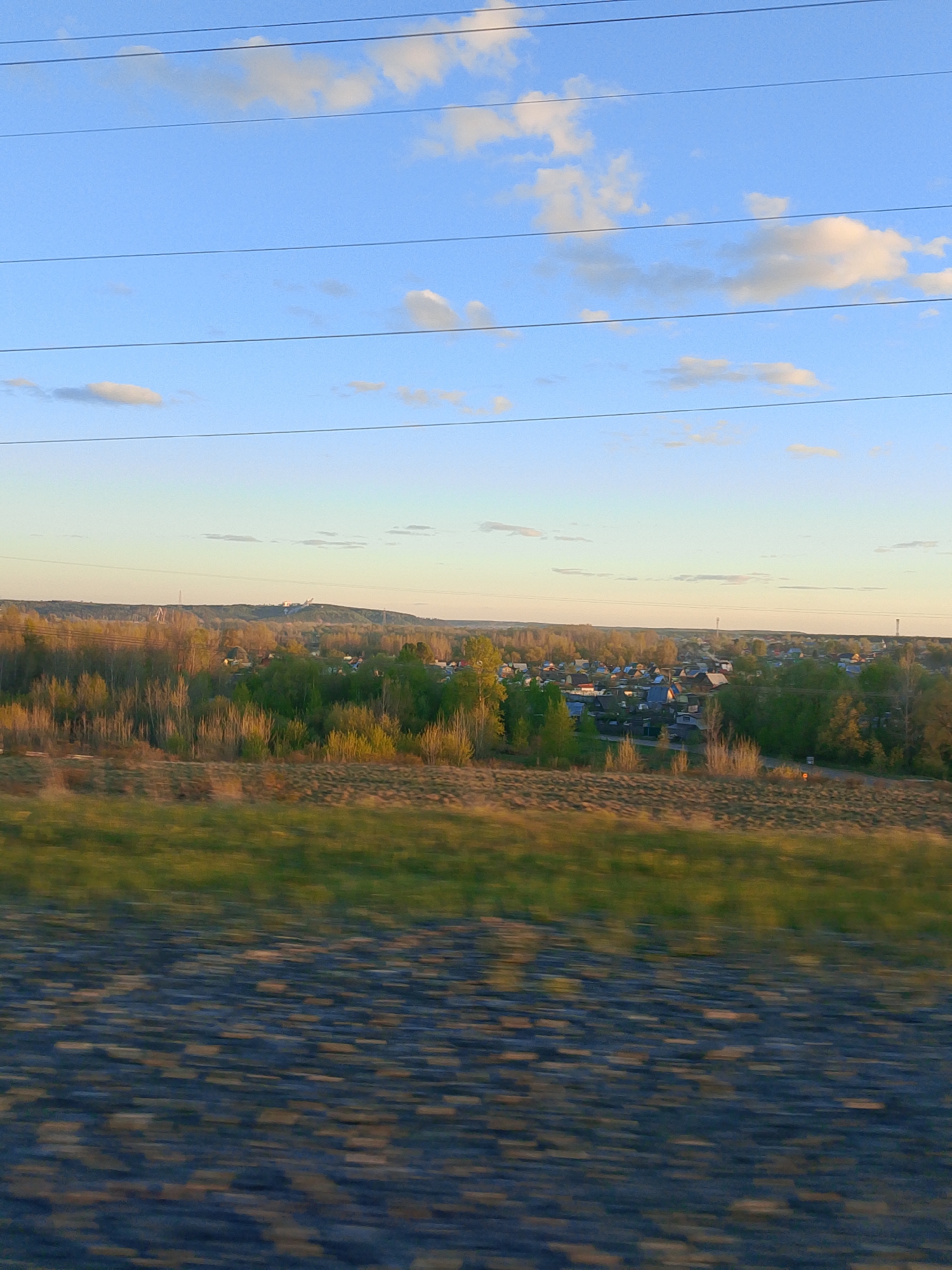 The image shows a landscape in the city of Chaikovsky, Russia. The sky is blue with a few scattered white clouds. Power lines stretch across the upper portion of the image. In the middle ground, there's a mix of trees and shrubs with the tops of some houses visible, suggesting a residential area. In the far background, there appear to be industrial structures or buildings on a hill. The foreground shows blurry grass and a surface that looks like road or track ballast, suggesting the photo was taken from a moving vehicle.
