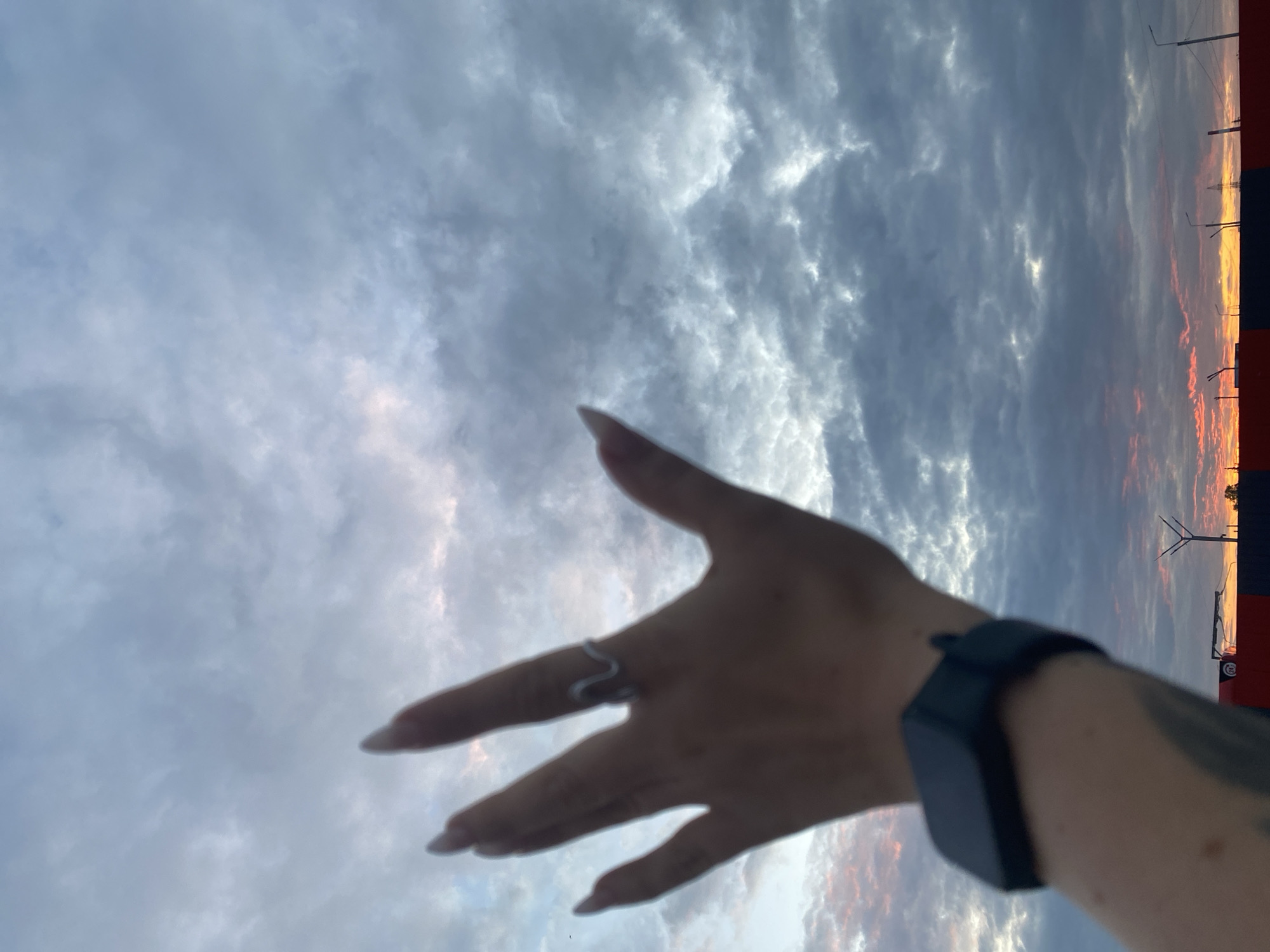 The image captures a close-up of a person's hand, adorned with a ring and a smartwatch, extended towards a cloudy sky. The sky is filled with dramatic clouds, with hints of pink and orange hues visible near the horizon, suggesting either a sunrise or sunset. In the background, a red and black structure runs horizontally across the frame, with streetlights and what appear to be industrial elements visible above it. The presence of a road sign with the number "24" on it is also noticeable. The scene is likely situated in an outdoor urban environment, possibly on the outskirts of Neftekamsk, Russia, given the context of the prompt. The overall mood is one of contemplation or reaching out towards the sky.