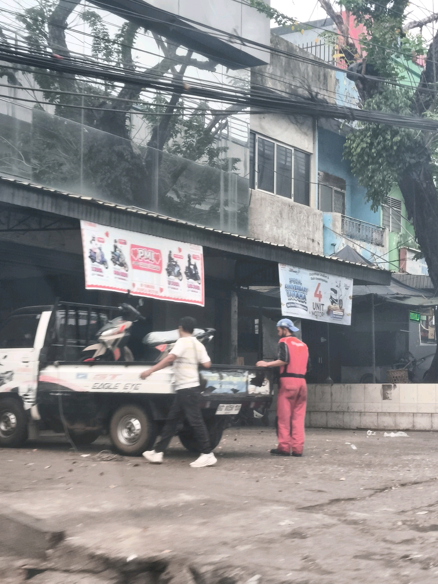 This image depicts a street scene in Palembang, Indonesia. In the foreground, a white pickup truck is parked, its bed carrying at least two motorcycles. Two individuals are present. One, wearing a red vest and red pants with a blue cap, stands near the back of the truck, appearing to be engaged in some activity. The second person, wearing a light-colored shirt and dark pants, walks away from the truck towards the left side of the frame. Above the truck, a banner is hung, displaying images of motorcycles and text that appears to be promotional. The background shows a multi-story building with windows and balconies, and trees with dense foliage. The overall environment suggests an urban setting, possibly a commercial area, with overhead power lines and a slightly weathered appearance to the buildings.
