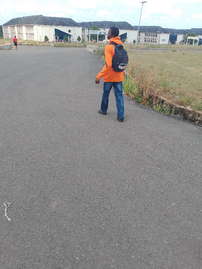 The image shows a person walking on a paved road in Umueshi, Nigeria. The individual is wearing an orange hoodie, blue jeans, and a black backpack. They are walking away from the camera. In the background are several institutional buildings. A second person is further away to the left. The environment is characterized by a paved road, grassy areas, and various buildings.