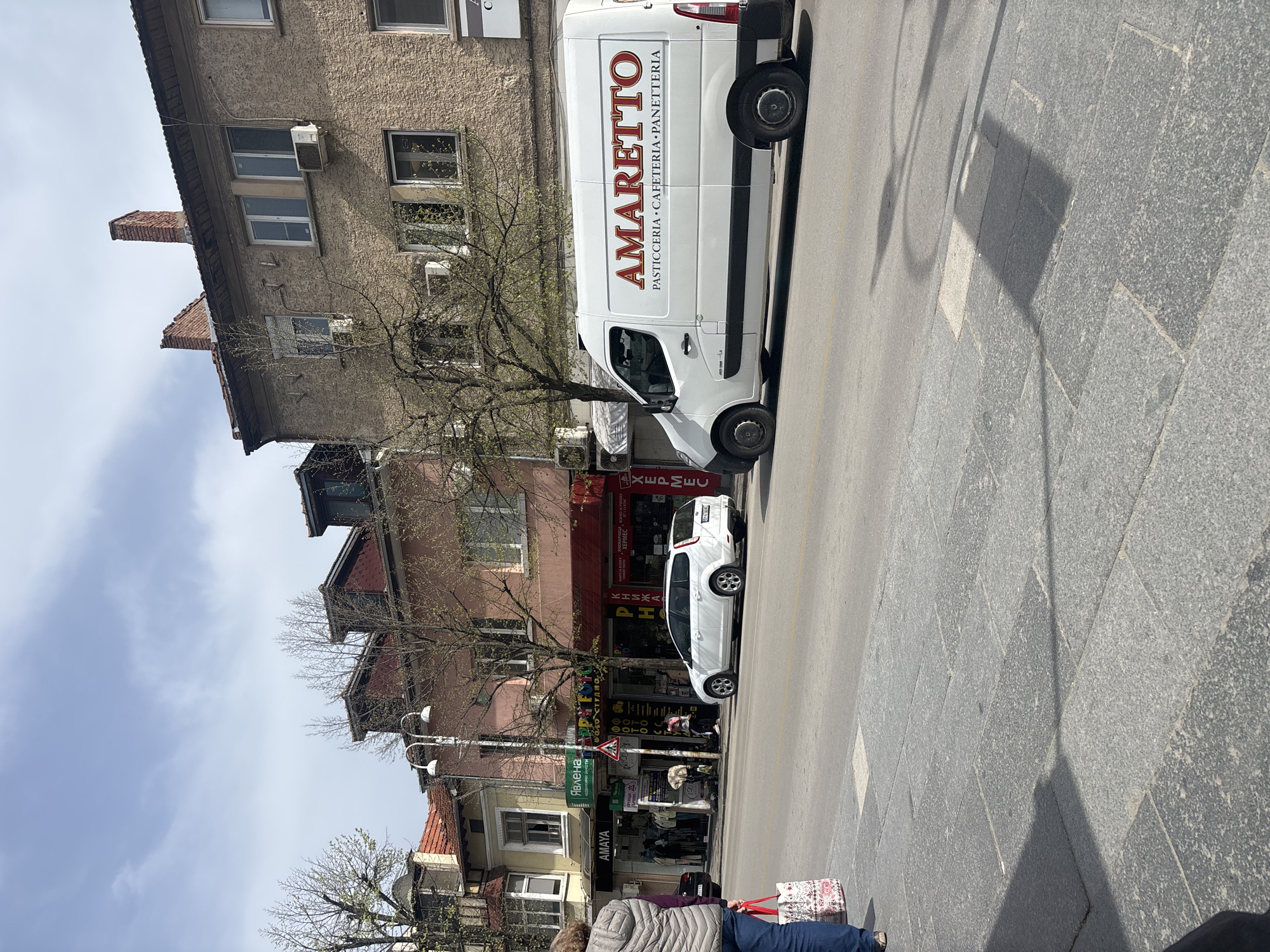 The image depicts a street scene in Veliko Tarnovo, Bulgaria. An "Amaretto" delivery van, a white hatchback car, and pedestrians populate the area. Buildings with varied architectural styles line the street. Signs indicate local businesses. A pedestrian walks on the sidewalk. The sky is partly cloudy.