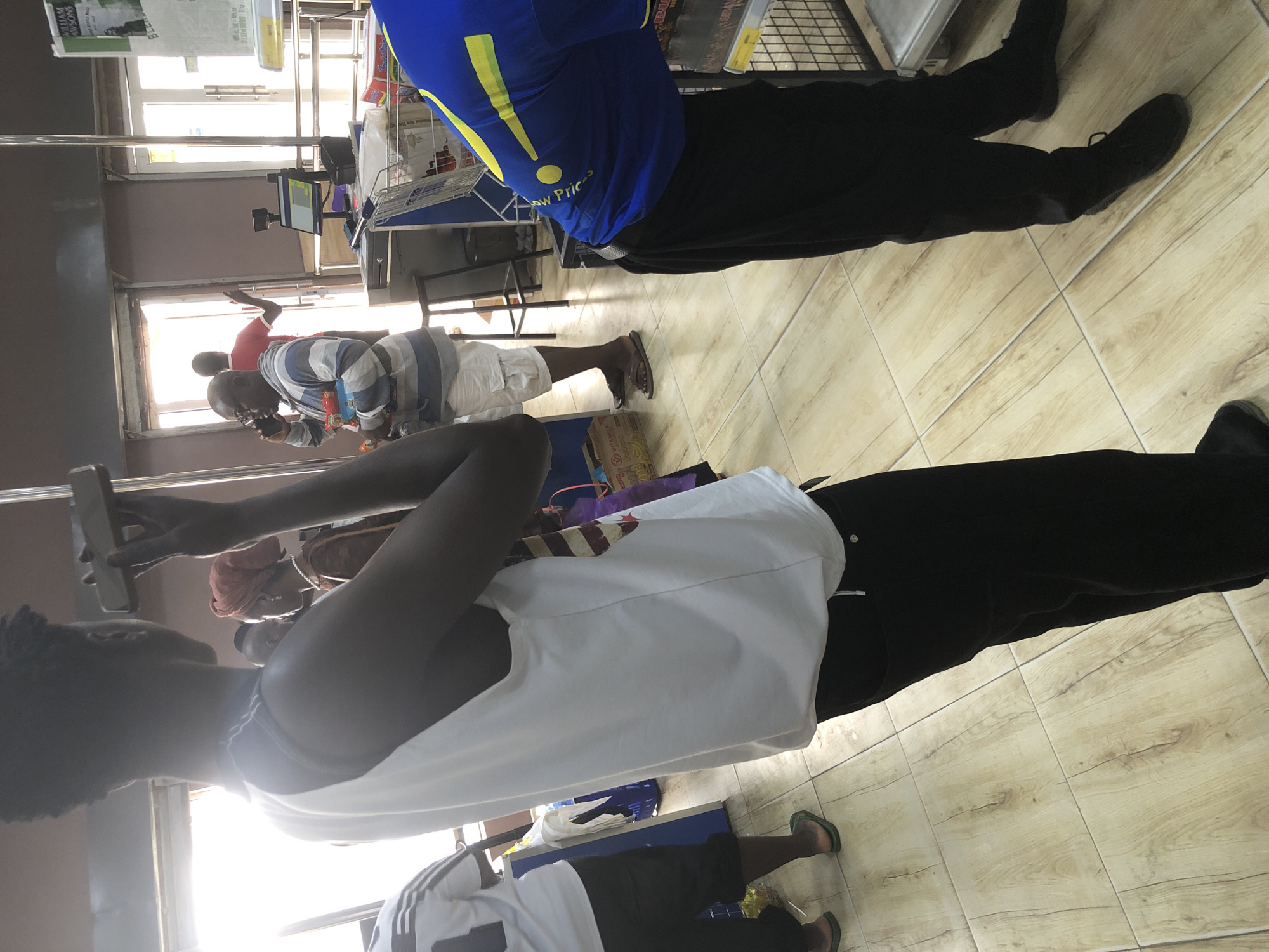 The image depicts a scene inside what appears to be a retail store, possibly a supermarket in Ado, Nigeria. In the foreground, a person in a white tank top and black pants stands with their back to the camera, holding a mobile phone up to their ear. Their right hand is extended, and they are looking towards the side. To the left, another person wearing a white t-shirt with black stripes on the sleeves and black shorts is visible from the waist down, walking away from the camera and wearing green flip-flops.
In the background, a man wearing a striped shirt and white shorts is on his phone and holding several items. To his right, a person wearing a red headscarf and a brown garment is also visible. Behind them, a doorway leads to an outdoor area with bright natural light. Further to the right, a person in a blue shirt with yellow accents is bending over, looking at something on a shelf. A shopping cart and various products are also visible in the store environment. The floor is tiled with a wood-like pattern. The overall impression is a busy, everyday shopping scenario.