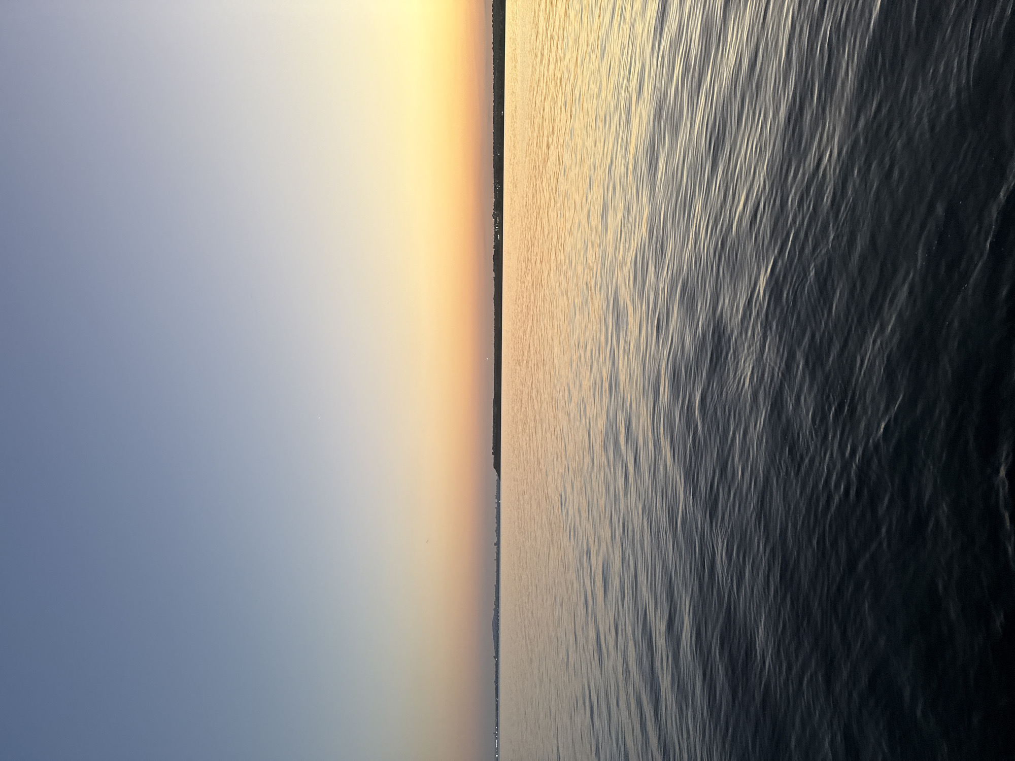 The image captures a serene seascape at dusk, likely along the coast of Pomorie, Bulgaria. The sky transitions from a soft blue at the top to a warm gradient of yellow and orange near the horizon, indicating the setting sun. The sea is calm, with gentle ripples reflecting the colors of the sky. In the distance, a faint silhouette of a city skyline is visible along the coast, with some buildings appearing as small lights. The foreground is dominated by the dark, textured surface of the water, with subtle highlights from the sky's illumination. There are no people, animals, or specific objects prominently featured in the scene, making it a purely environmental and atmospheric depiction.