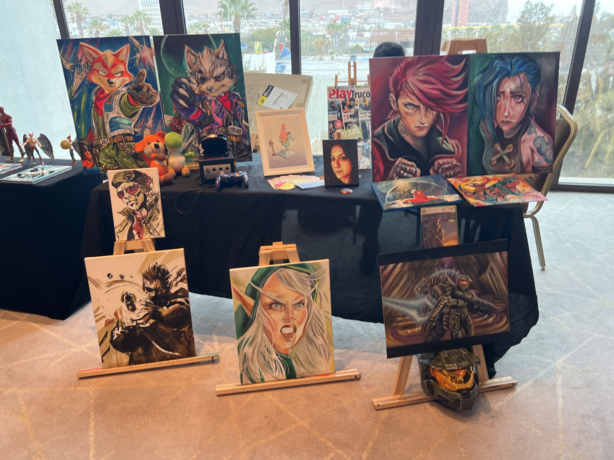This is a photograph taken at an event in Arica, Chile, featuring an artist's display table filled with various paintings and merchandise. The table is set up indoors, likely in a convention center or hotel, with a large window in the background offering a view of a city skyline and palm trees.
The display includes several framed and unframed paintings. Prominently featured are two large portraits of female characters with distinctive hairstyles and expressions, one with red hair and the other with blue hair. To the left of these are two larger paintings of anthropomorphic animals, one resembling a red fox and the other a grey wolf, rendered in a vibrant, stylized manner. Smaller paintings and framed artwork depict characters such as Solid Snake from Metal Gear Solid, an elf-like character with a fierce expression, and a scene from the Halo video game series, complete with a Master Chief helmet placed in front of it. There are also plush toys, video game controllers, and magazines on display.
Several individuals are visible in the background, one of whom appears to be browsing the artwork. The lighting suggests it is daytime. The overall scene conveys a lively atmosphere of an art exhibition or fan convention.