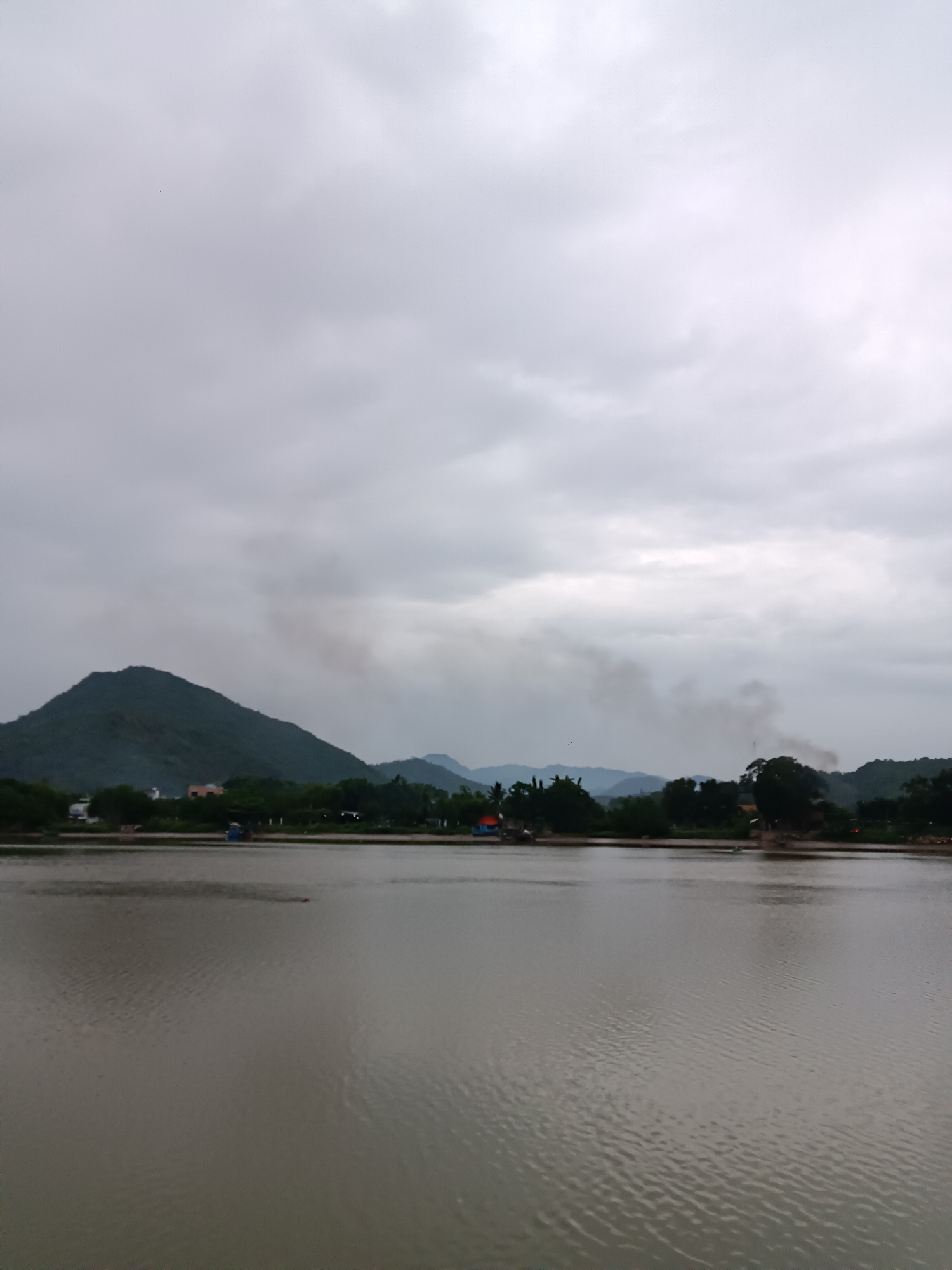 The image depicts a serene body of water with a treelined shore and distant hills under a cloudy sky. In the foreground, the water is calm, reflecting the muted light. The middle ground shows a strip of land with lush green trees and some scattered buildings, suggesting a semi-rural or park-like setting. A large, rounded hill dominates the left side of the background, with several smaller, layered hills extending to the right. Wisps of dark smoke rise from behind the trees in the mid-ground, possibly from a distant fire or industrial activity. The overall atmosphere is subdued due to the overcast weather. It is highly probable that this scene is located in the city of Nha Trang, Vietnam, given the typical landscape of rolling green hills and bodies of water found in and around the city.