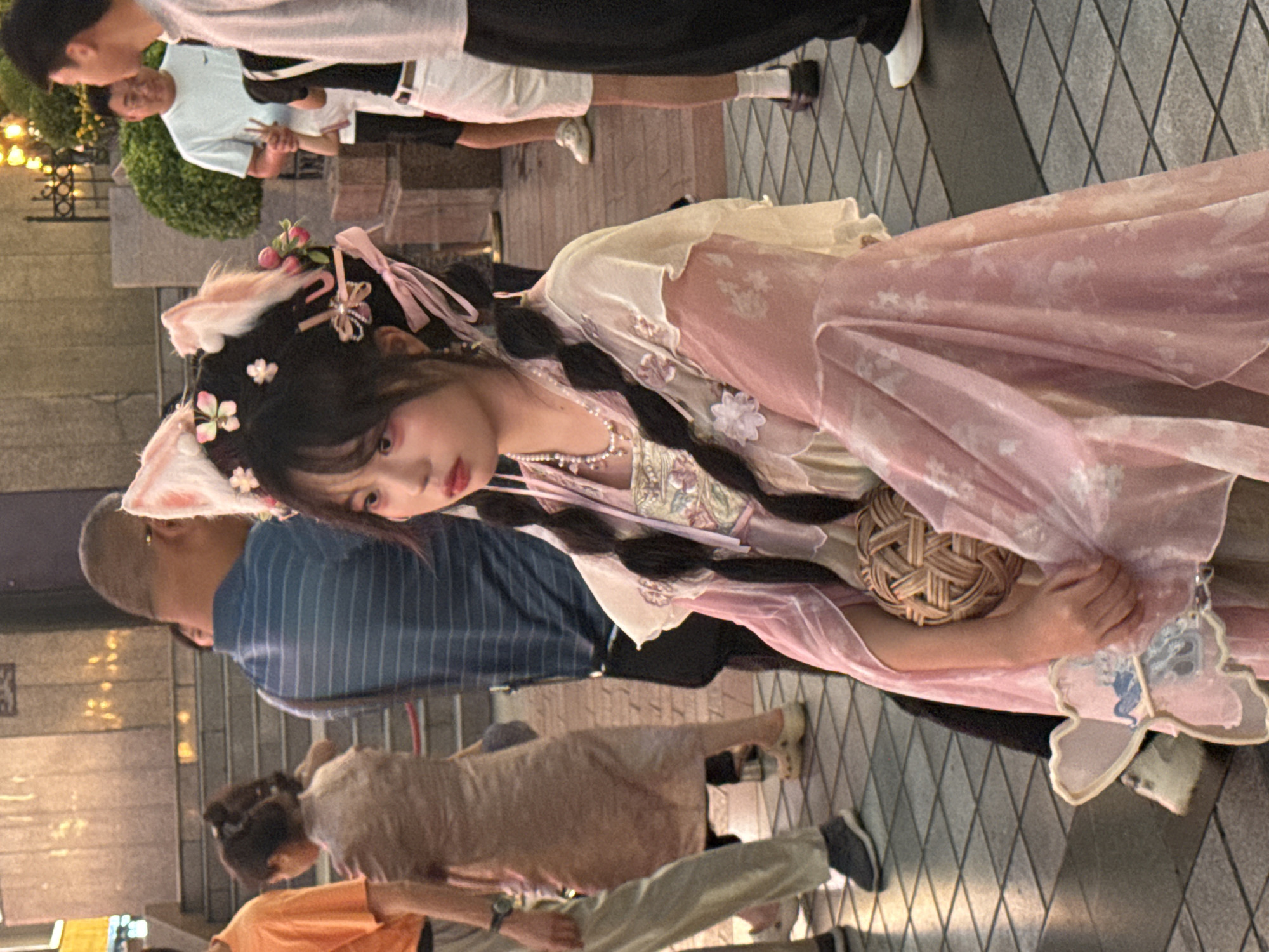 In the city of Yuzhong District, China, a young woman dressed in a pink and white traditional Chinese outfit, possibly a hanfu, is the central subject. She is adorned with cat ears and floral hair accessories, with her dark hair styled in two braids. She wears a pearl necklace and holds a woven ball in her right hand. A pink fan is visible near her left hand. The background shows a crowd of people, some walking and some standing. The environment appears to be an outdoor urban setting with tiled ground and a building with stairs in the background, suggesting a public space such as a plaza or street. The lighting indicates it might be evening or dusk.