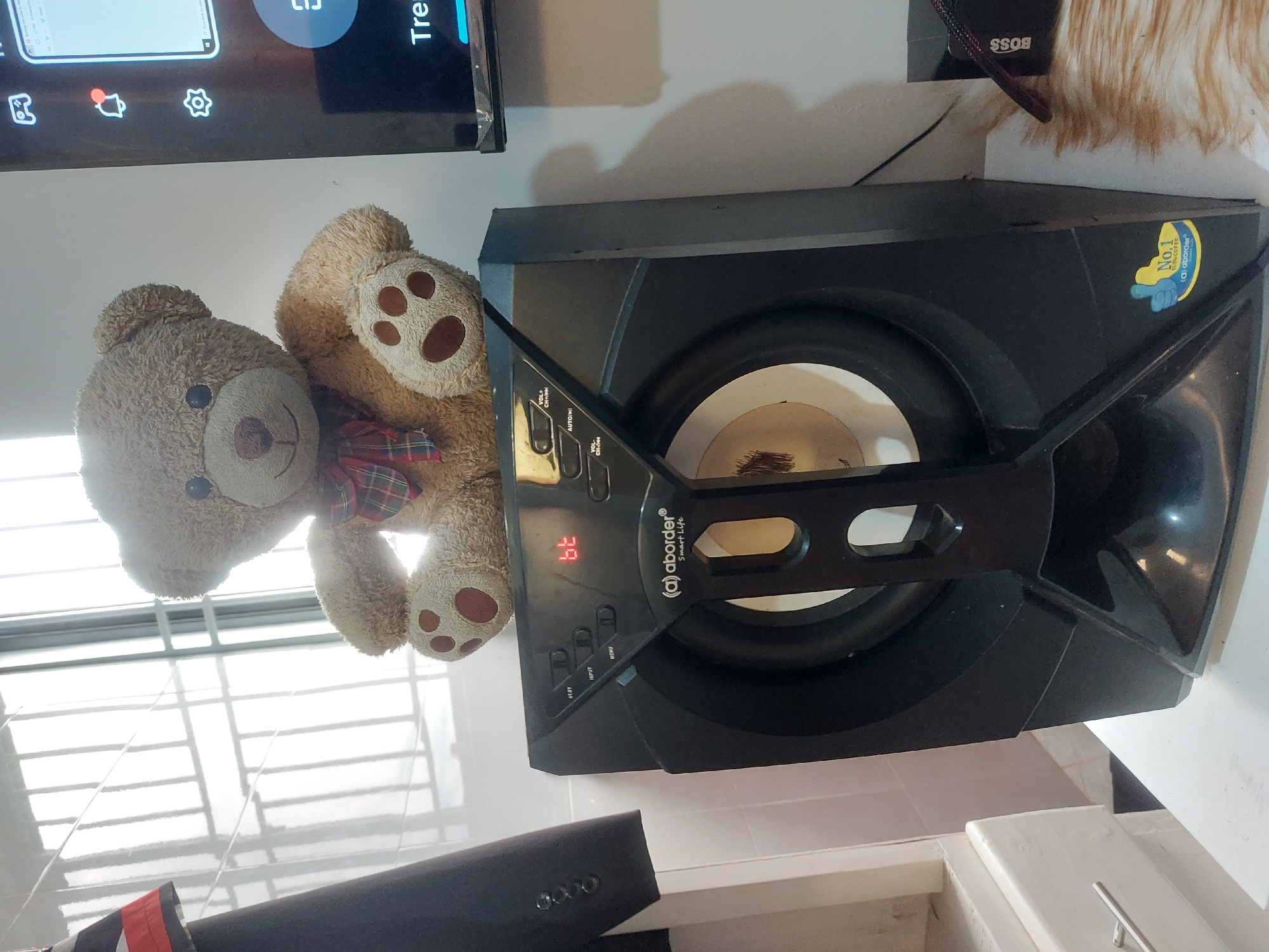 A teddy bear wearing a plaid bow tie is propped against a black subwoofer bearing the brand name "aborder". The display panel on the subwoofer shows the number "39". A section of a TV screen is visible in the upper left corner of the image. The environment appears to be indoors, with the presence of window blinds. A black object is visible at the bottom of the frame. No information is discernible in the image as to the location being the city of Dar es-Salaam, Tanzania.