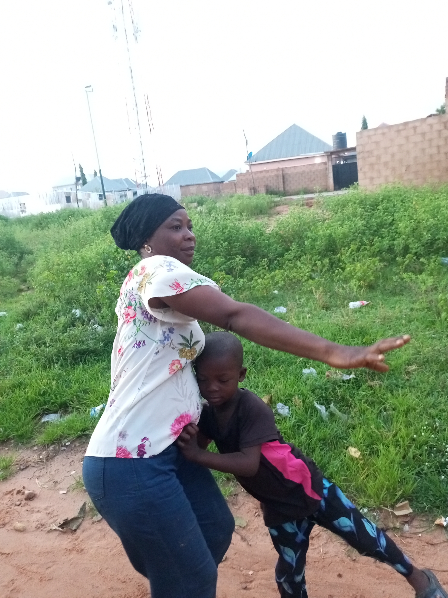 In Tempure, Nigeria, a woman wearing a black headscarf and a floral print shirt is embracing a young child. The child is wearing a black t-shirt with a pink accent and patterned leggings. They are standing on a dirt path with lush green vegetation surrounding them. In the background, there are buildings, including a structure with a light blue, triangular roof and a brick wall. A tall communications tower is also visible in the distance against an overcast sky.