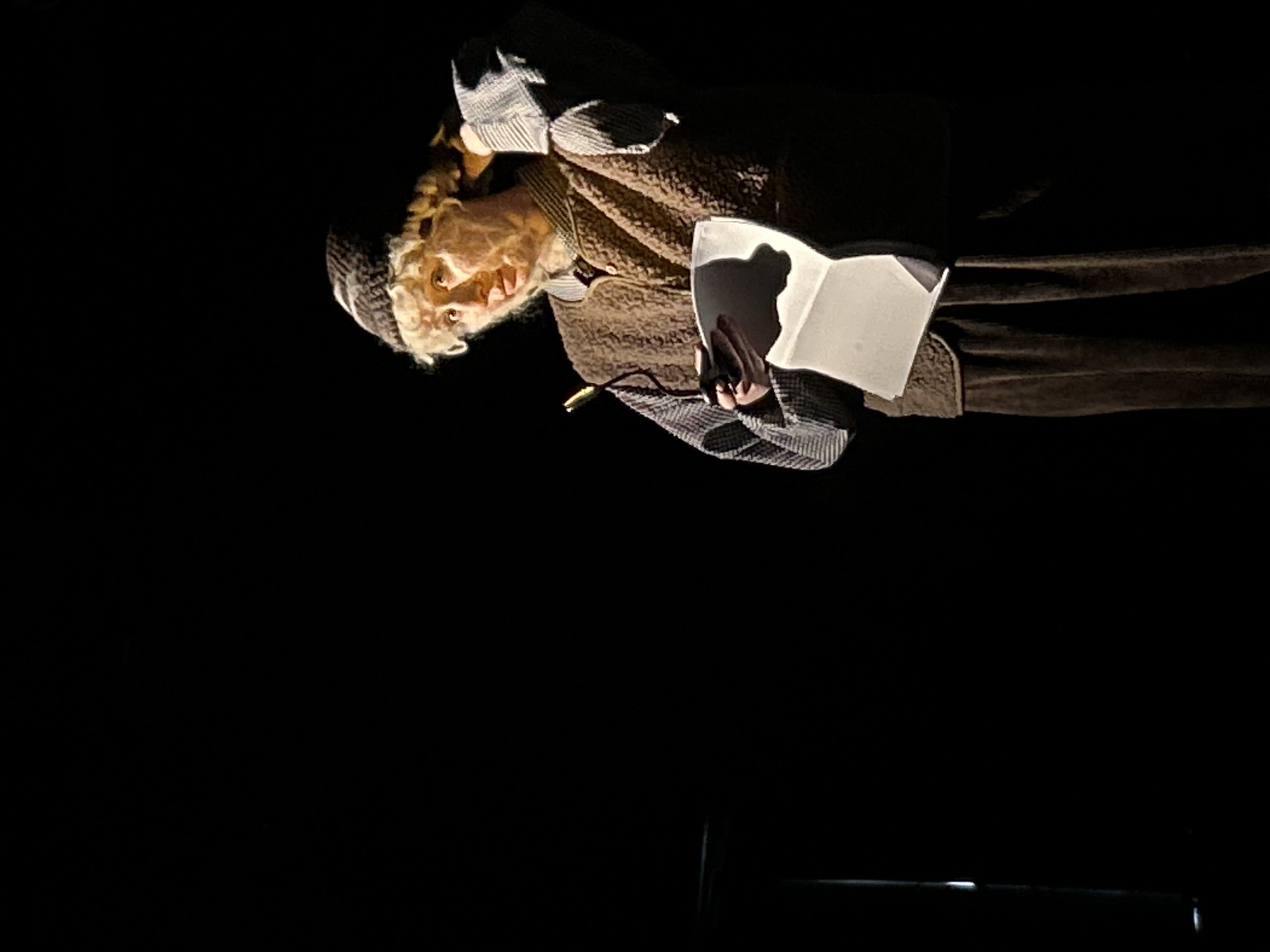 An individual wearing a hat, coat, and gloves is holding an open book in a dark environment. The person's face is illuminated by a light source, highlighting their features. A dark horizontal bar is visible in the lower portion of the frame. The setting is unknown.
