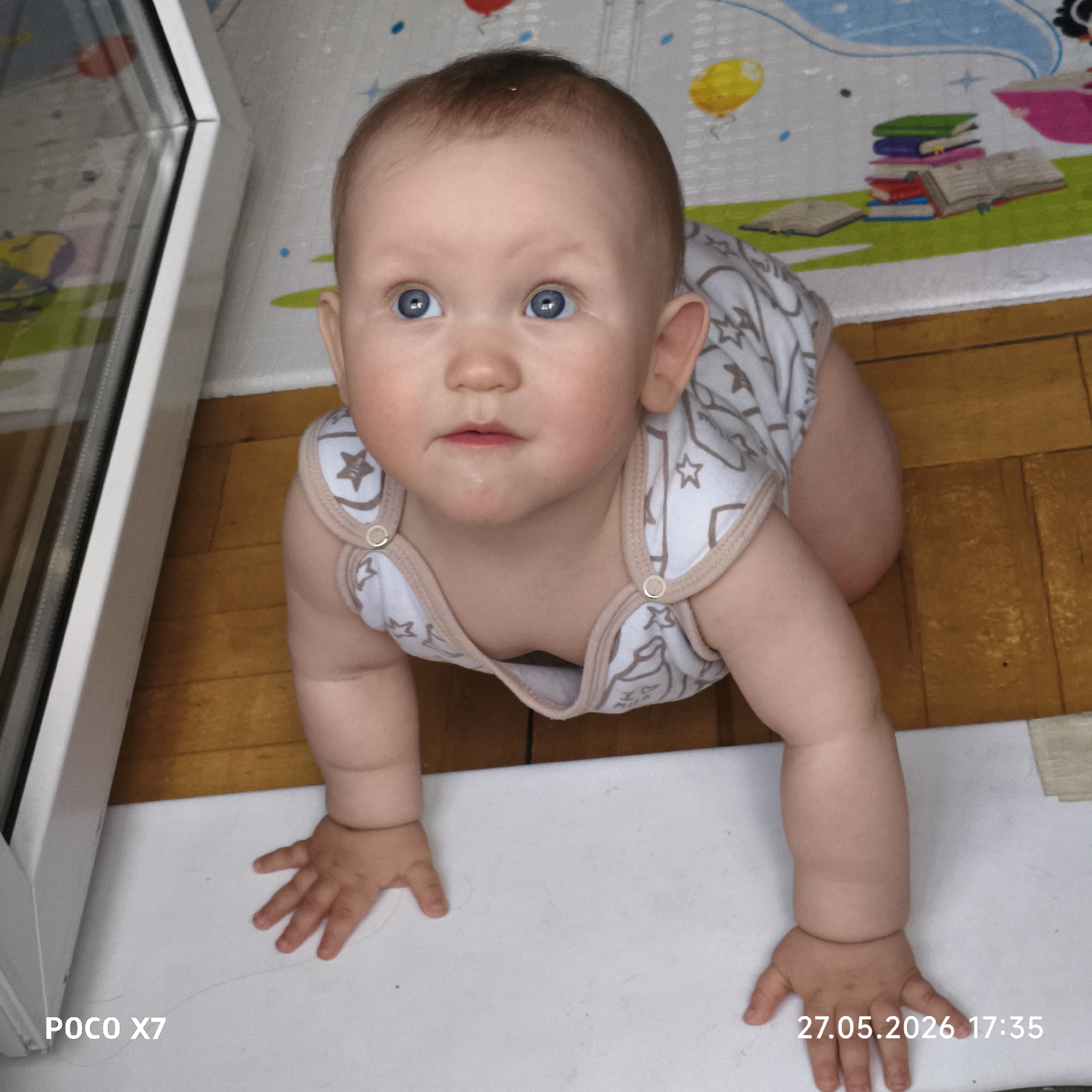 A baby with blue eyes is crawling on a wooden floor, next to a white window frame. The baby is wearing a white and gray onesie with star patterns. A play mat with a stack of books is visible in the background. The image was taken with a POCO X7 and includes a timestamp of 27.05.2026 17:35. Location is unknown.
