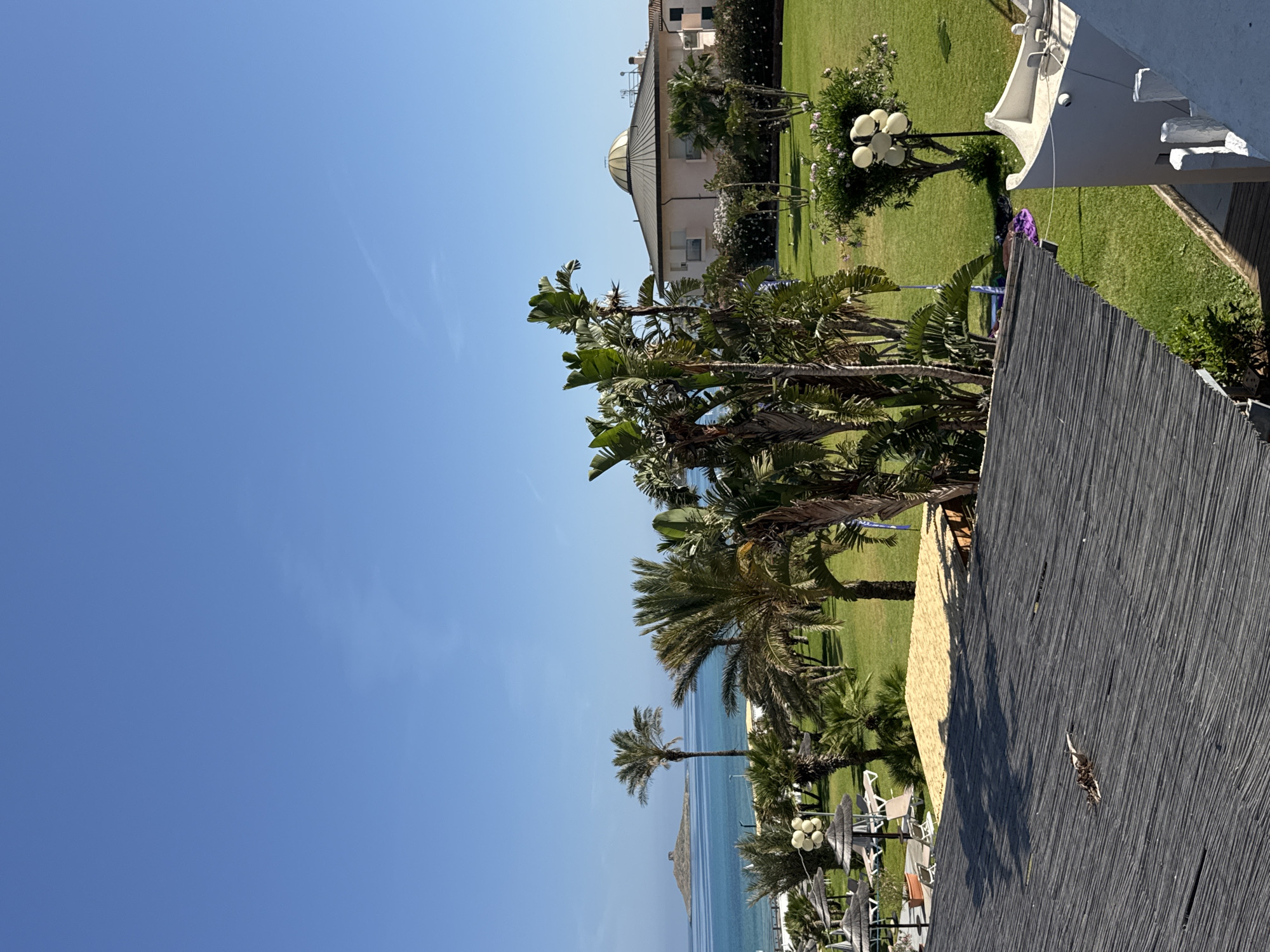 The image depicts a sunlit coastal resort in Capaci, Italy, on a clear day. The sky is bright blue, with a faint, wispy contrail visible in the upper mid-right. In the distance, a tranquil turquoise sea stretches to the horizon, featuring a small, rocky island with a prominent, ancient-looking stone structure (possibly a tower or ruin) situated upon it.
The midground is dominated by an expansive, well-maintained green lawn interspersed with numerous palm trees and large-leaved tropical plants. Within this landscaped area, several thatched sun umbrellas and sun loungers are arranged on what appears to be a paved or decked surface. Towards the right, a distinctive, light-colored, circular building with a large, shallow, domed roof is visible, surrounded by lush green foliage and flowering shrubs. A multi-globe outdoor lamp post stands on the lawn to the right.
The foreground presents a view from an elevated position, looking down onto a dark, textured roof, possibly constructed from interwoven reeds or bamboo slats. Scattered debris, including dried leaves and small twigs, rests on this roof surface. To the immediate right, the white upper section and architectural details of an adjacent building are partially visible. Below the foreground roof, a section of lower green lawn shows several pieces of colored fabric, including a prominent purple item, lying on the grass. No human figures are clearly discernible, but the presence of loungers and fabrics suggests human activity.
